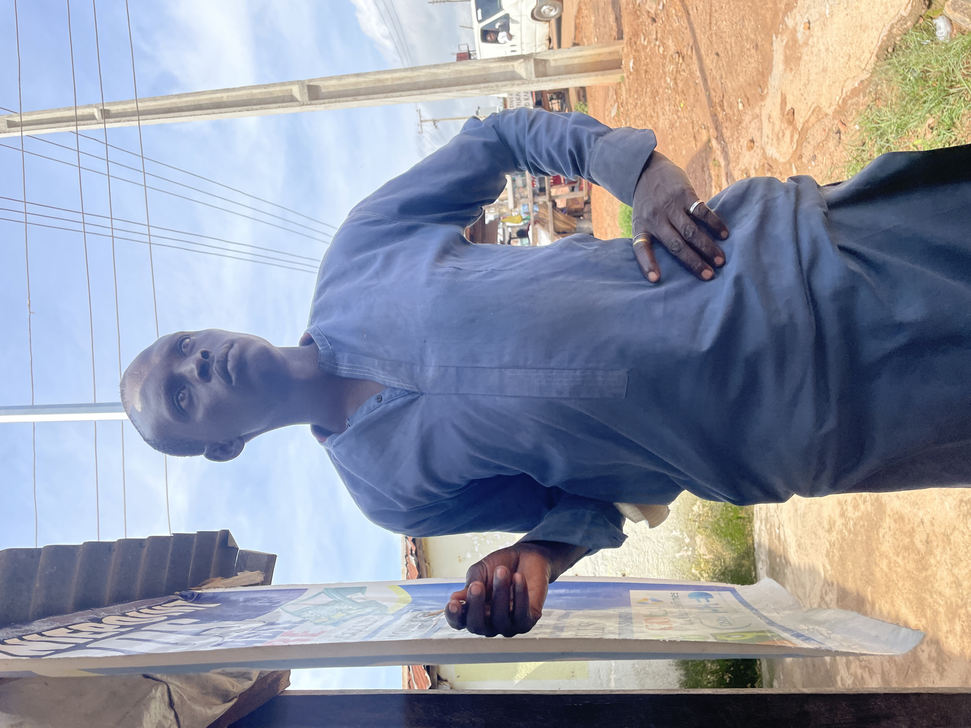 A man stands outdoors in Jos, Nigeria, looking to his right. He is wearing a blue, long-sleeved garment with a band of intricate stitching around the collar and down the front placket. His left hand is at his hip, adorned with two rings, while his right hand is held up near his face, with his index finger and thumb lightly touching. He has dark skin and is bald.
The man is positioned in front of a stall or shop with a corrugated metal roof and a large, blue and white banner advertising various products or services. To his right, a concrete utility pole stands tall, with numerous power lines crisscrossing the sky. Beyond the man and the pole, the ground is a reddish-brown earth, with patches of dry grass. In the background, a white van is parked, with a person visible through the open window. Other structures, possibly market stalls or houses with tin roofs, are visible further in the distance. The sky is cloudy, suggesting an overcast day. The lighting is diffused, characteristic of an outdoor environment.