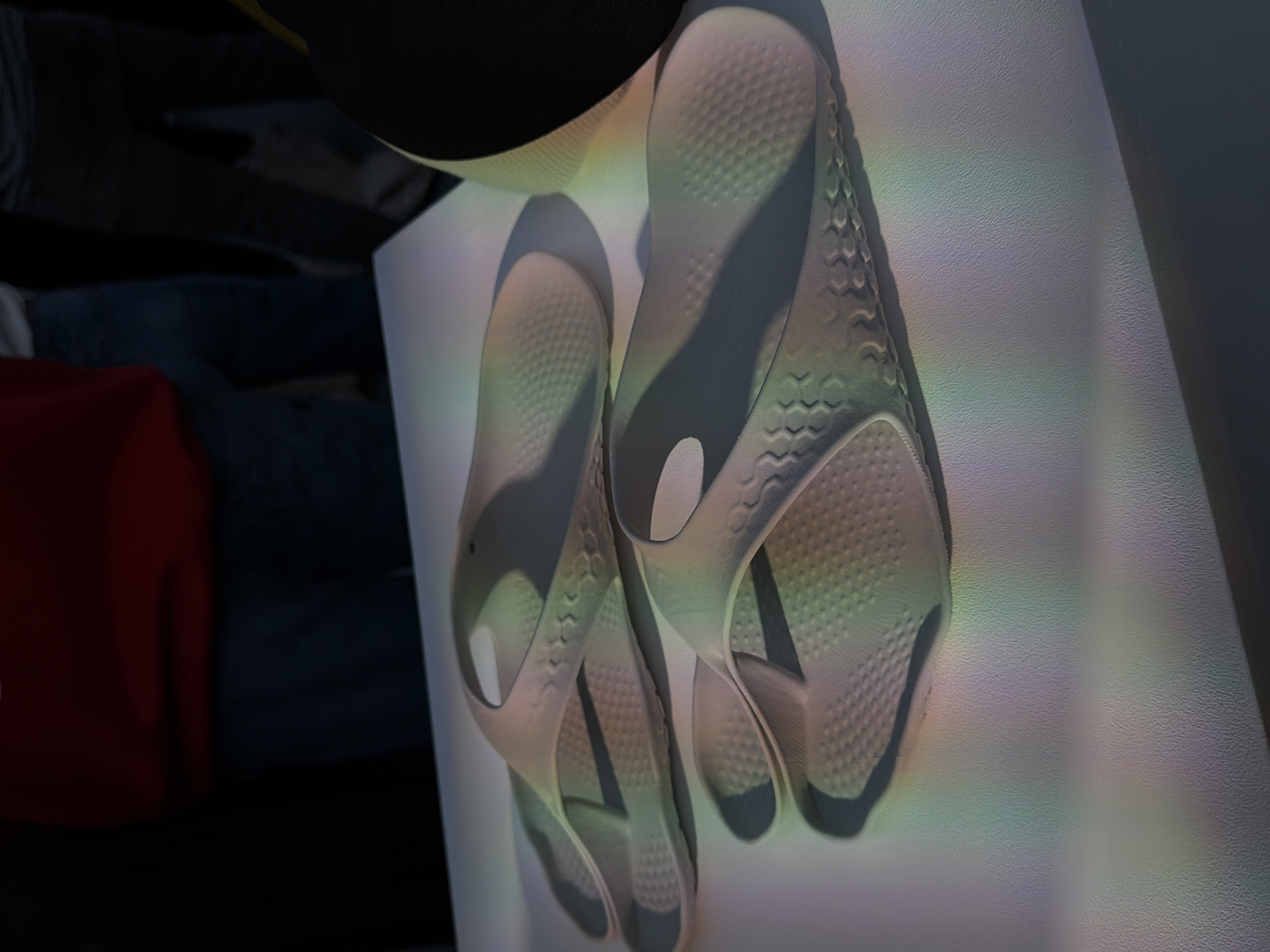 The image presents a pair of gray sandals placed on a white surface, possibly a table or platform. The sandals are of a modern design, featuring a honeycomb pattern on the sole. The lighting in the scene is soft and diffused, with rainbow-like colors casting across the sandals and the white surface. The background is somewhat blurred, showing glimpses of a red object and what appear to be denim jeans, indicating an indoor setting. There's a portion of a black object, possibly clothing, visible at the top. The overall impression is a stylized product shot, potentially for advertising or aesthetic purposes. The image does not show any identifiable location in London, United Kingdom, nor does it depict any specific actions, interactions, or people.