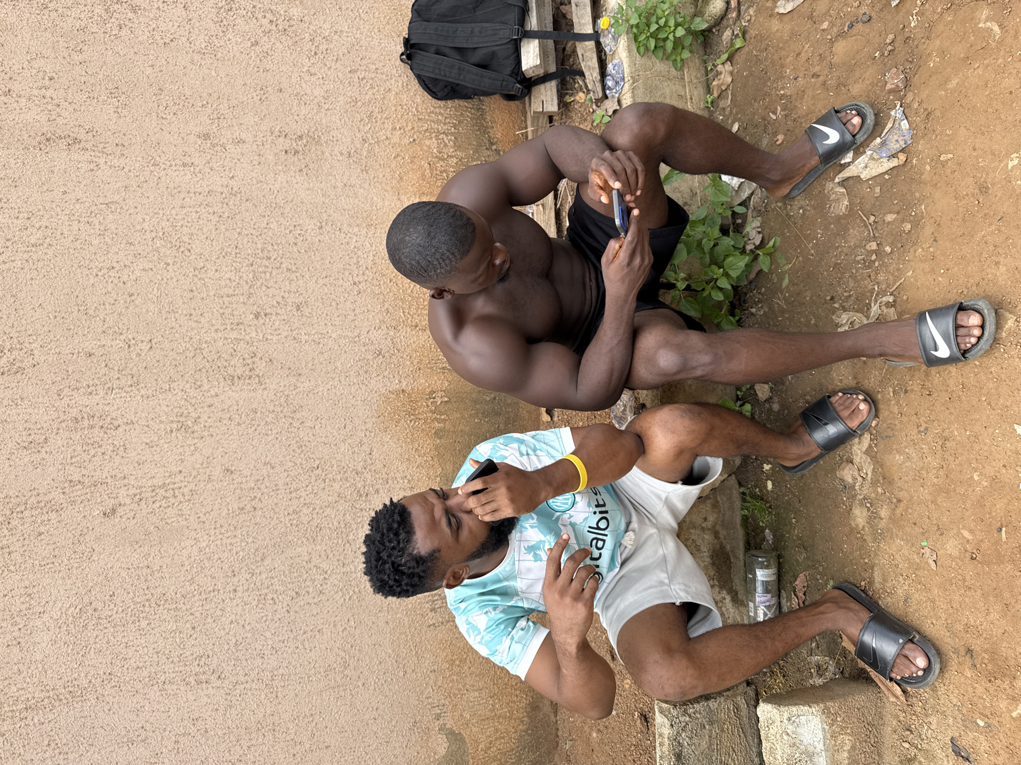 Two Black men are sitting outdoors in Akwanga, Nigeria, near a textured, beige wall. The man on the left, wearing a light blue and white patterned t-shirt and light grey shorts, is holding a black smartphone to his ear with his right hand and touching his nose with his left. He has a yellow wristband on his left wrist. The man on the right, shirtless and wearing black shorts, is looking down at a smartphone in his hands. A black backpack is placed on some wooden planks behind him. The ground is dirt and there is some sparse green vegetation. An empty glass bottle lies on the ground between the two men. Both men are wearing black flip-flops.