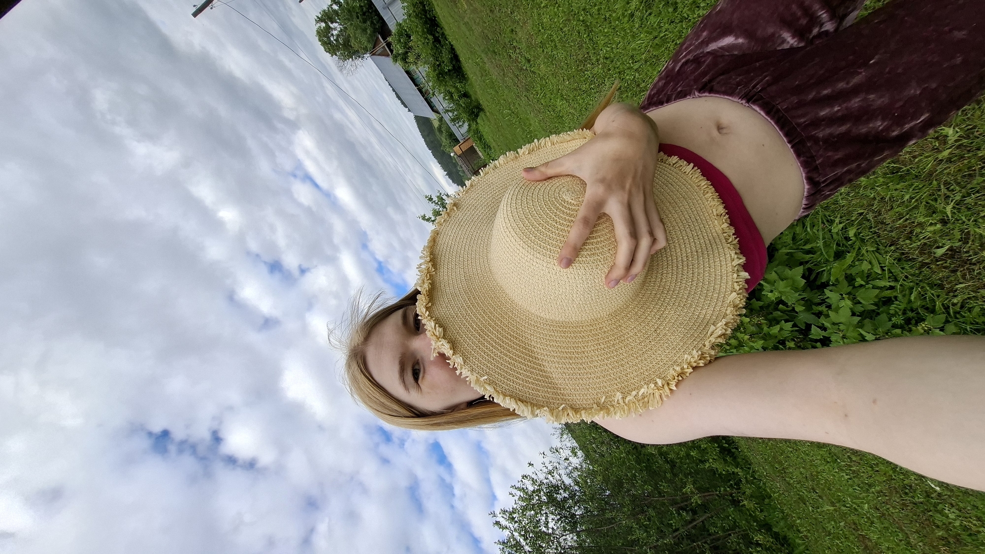 A young woman with long blonde hair is holding a wide-brimmed straw hat in front of her face, obscuring her lower face. She is wearing a pink top and dark velvet pants. She is standing in a grassy area with green bushes and trees behind her. In the background, there is a wooden fence, a house, and a hill. The sky is partly cloudy. The image was taken in Большой Букор, Russia.
