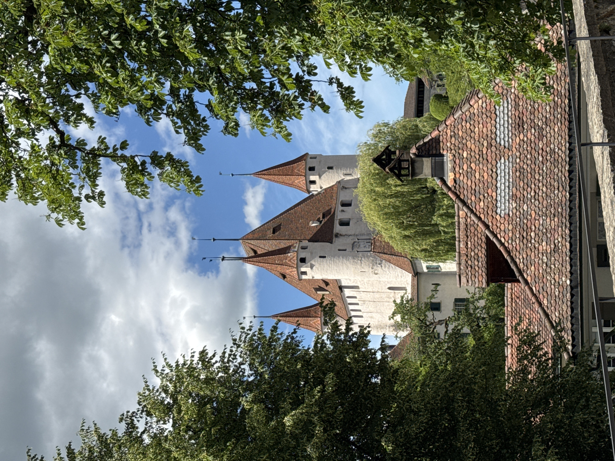 This media file shows a view of the Thun Castle in Thun, Switzerland. The castle has multiple white towers with red tiled roofs and pointed spires. The perspective is from below, looking up towards the castle, framed by lush green trees. In the foreground, there are also roofs of other buildings with red tiles. The sky is blue with scattered white clouds, suggesting a pleasant day. The image is oriented vertically, with the trees appearing at the top and bottom, and the castle in the middle. There is no visible text in the image. The scene appears to be a tourist attraction, with the castle likely being the main subject of interest. The time of day appears to be daytime, given the bright lighting. The weather is fair and sunny.