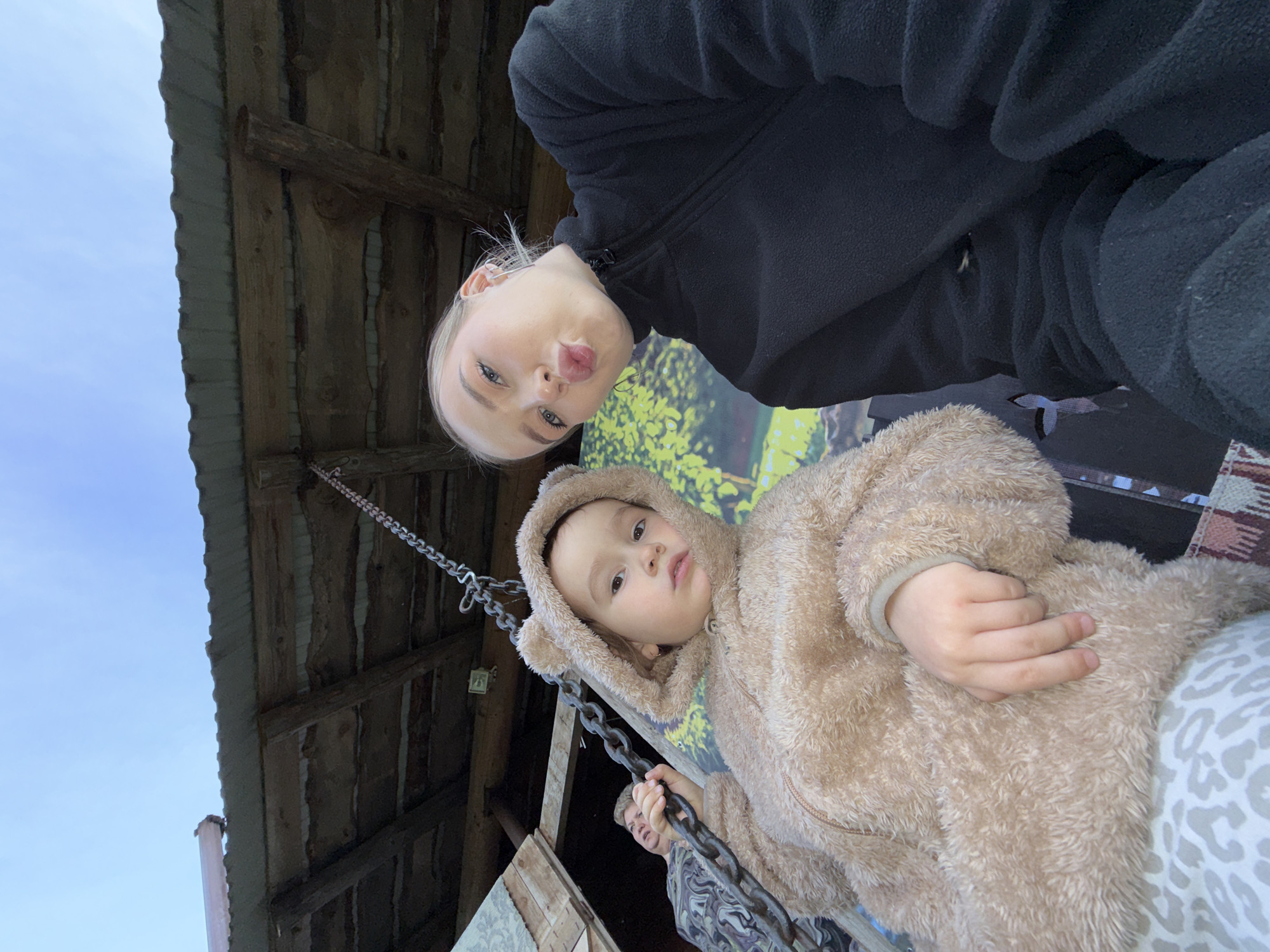 This low-angle, tilted photograph captures two individuals looking down towards the camera, beneath a rustic wooden structure, against a backdrop of blue sky and blurred greenery.
The primary subjects are an adult and a young child. In the upper portion of the frame, an adult with blonde hair, light-colored eyes, and dark eyebrows wears a black fleece jacket and makes a playful, pouting expression towards the viewer. Below and slightly to the right of the adult is a young child, appearing to be a toddler or preschooler. The child is dressed in a cozy, light brown, fluffy hooded jumpsuit or jacket that features small ear-like details on the hood, resembling an animal costume. The child looks directly at the camera with a neutral, slightly curious expression, holding onto a heavy metal chain with both hands. A third person's head and shoulder, wearing a patterned top, are partially visible behind the child's right side.
The setting appears to be outdoors during daytime under a clear sky, as indicated by the bright blue patch visible on the left. Above the subjects is an overhead structure made of weathered wooden planks and beams, suggesting a shed, barn, or a covered porch. A light-colored plastic pipe is visible along the far left edge of the frame, attached to the wooden structure. Behind the subjects, a blurred area of green and yellow hues is visible, possibly foliage or a painted surface. A section of patterned fabric, resembling a rug or blanket, can be seen in the bottom right corner. The presence of the metal chains implies a swing or similar apparatus. The atmosphere is casual and playful, captured in what appears to be a rural or rustic outdoor location in Zhigalki, Russia. No visible text is present.