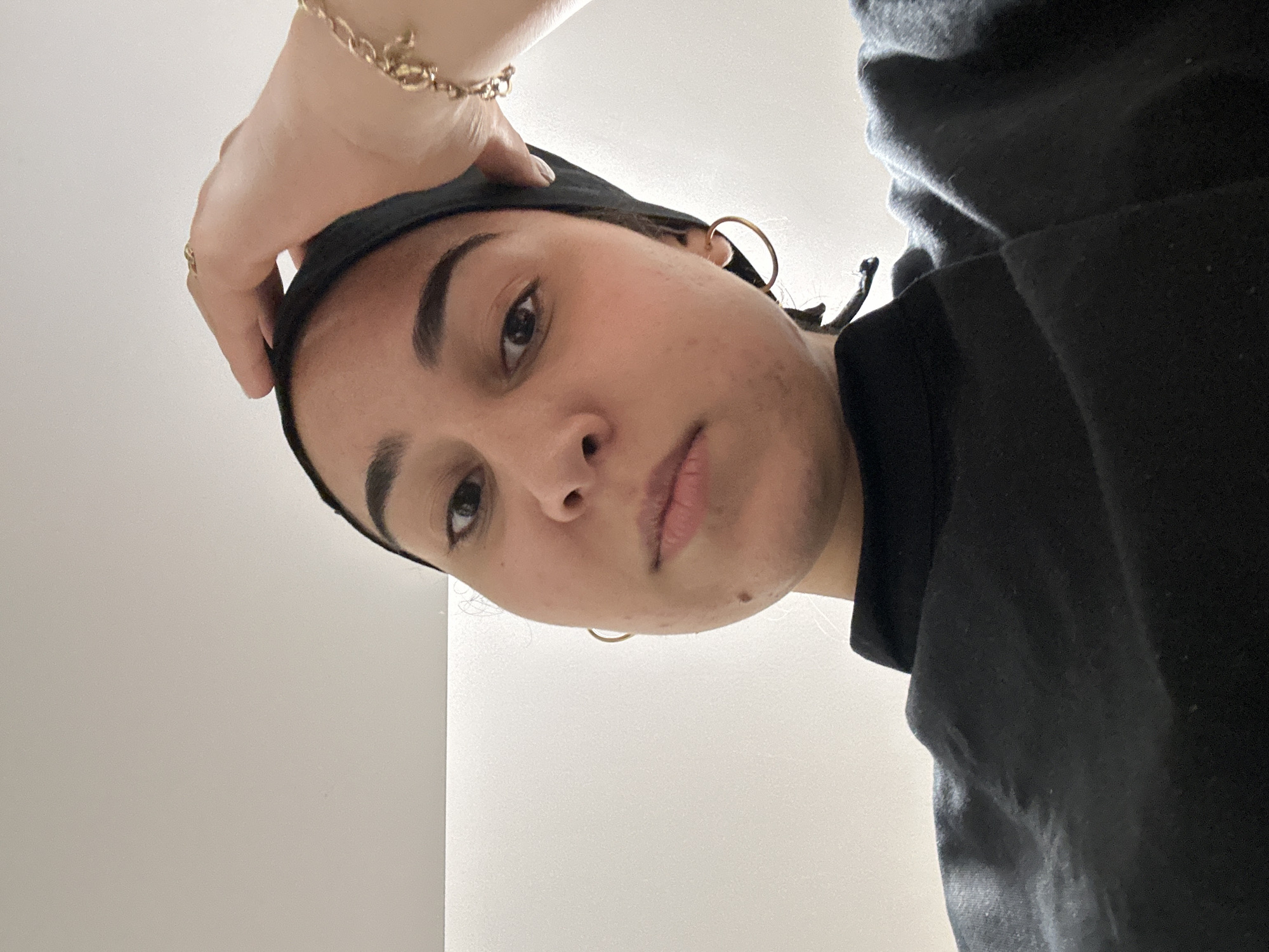 A woman with dark hair and eyebrows is looking down at the camera. She is wearing a black head covering and a black shirt. Her left hand, adorned with a gold bracelet, is on her head. She has gold hoop earrings in her ear. The background is a plain white wall. There is no visible information to identify the city of Burgas, Bulgaria.