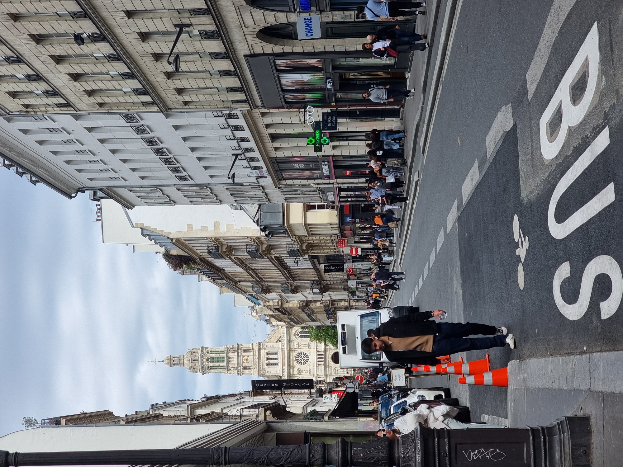 Here's a detailed description of the image:
**General Scene:**
The image is a street scene in Paris, France, specifically on a street with historical buildings and retail businesses. The photograph is taken at street level and is oriented vertically. A bus lane is prominent in the foreground.
**People:**
A few distinct groups of people are present:
* A man stands on the right side of the image, partially obstructing the bus lane. He is wearing a black jacket and dark pants and is holding a bottle in one hand.
* A group of people, likely pedestrians, are lined up near the storefronts of shops and businesses along the street.
* Further down the street are other individuals, likely walking or standing.
**Subjects and Objects:**
* **Buildings:** Tall, traditional Parisian buildings line both sides of the street. These buildings have multiple floors with windows, ornate architectural details, and balconies.
* **Église Saint-Vincent-de-Paul:** A large church with a prominent tower is visible in the distance.
* **Street Markings:** The street has marked bus lanes with the word "BUS" written in large white letters.
* **Retail:** Shops with signage such as "DUTY FREE", "CHANGE," and "nina kendosa" indicate the presence of retail businesses.
* **Traffic Control:** Orange cones are placed near the man standing in the foreground, possibly to indicate a construction zone or a temporary hazard.
* **Cars:** Some cars are parked along the street on the left side of the image.
* **Street Furniture:** A black decorative railing is in the foreground.
**Actions and Interactions:**
* People are walking, standing, and likely shopping along the street.
* The man is standing near the bus lane, interacting with the cones.
* The photographer is observing the scene from the street level.
**Environment:**
* The weather appears overcast or cloudy.
* The lighting is relatively muted.
* The street seems busy with pedestrian and vehicle traffic.