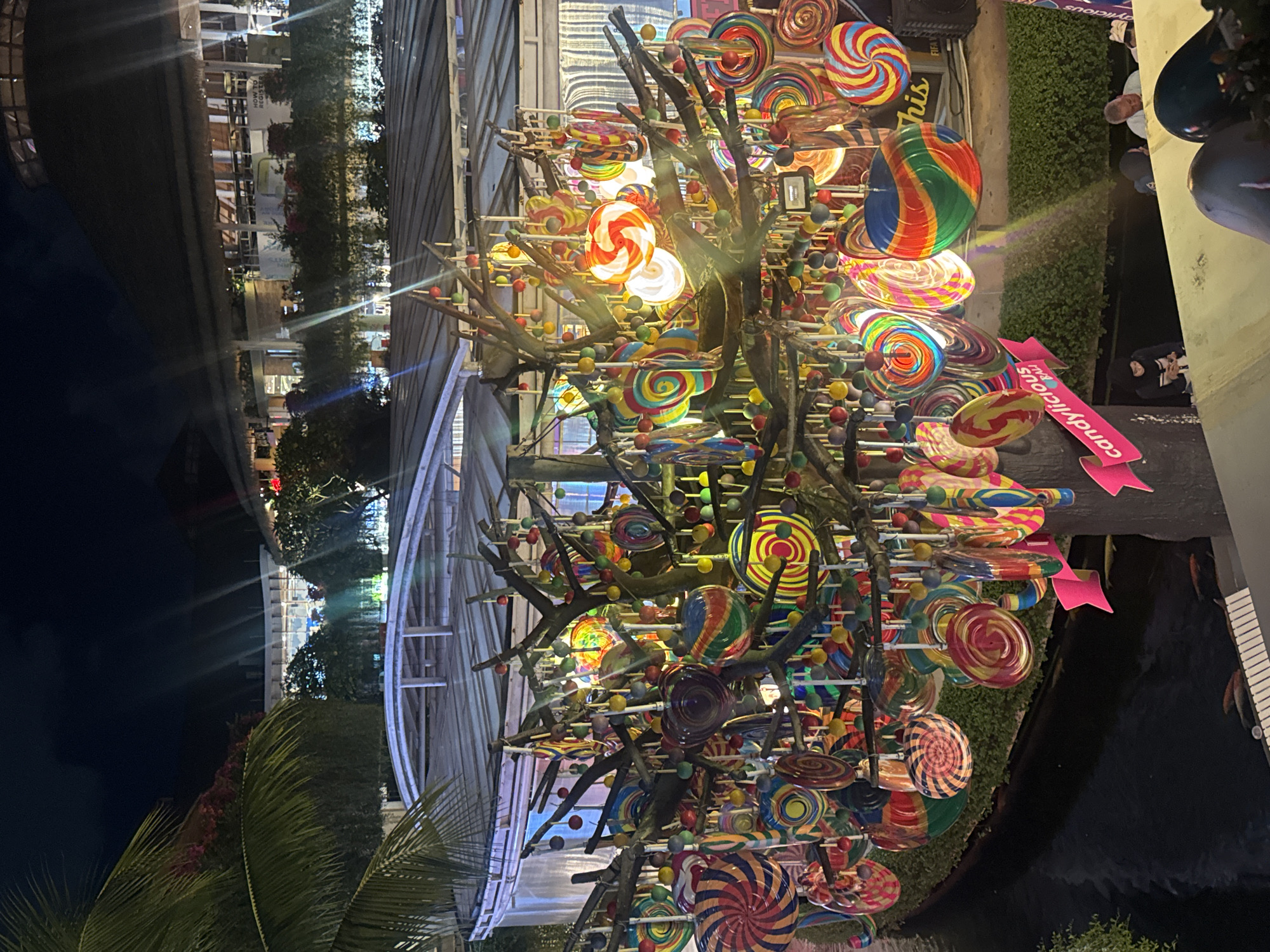 This image captures a vibrant display of colorful lollipops, arranged like fruit on the branches of a whimsical tree. The lollipops feature a variety of patterns and colors, including swirls, stripes, and solid hues. Interspersed among the lollipops are small, round candies in various bright colors. The display is illuminated from within, creating a warm and inviting glow that highlights the sugary creations. The lollipops are attached to thin branches that form a tree-like structure, some of which appear to be made of dark wood.
In the background, glimpses of a building with modern architecture can be seen, featuring glass windows and light-colored walls. There are also some lush green bushes and trees, suggesting an outdoor setting. The overall atmosphere is festive and cheerful, evoking a sense of childhood wonder and delight. The scene appears to be taking place at night, as indicated by the dark sky and the artificial lighting.
A pink ribbon with text is visible on the right side of the image. The text appears to read "candylicious." This suggests that the display might be part of a candy store or a special event related to sweets. There are also a few people visible in the background, some of whom are looking towards the lollipop display, perhaps admiring its artistry and vibrant colors. The presence of palm fronds in the foreground hints at a tropical location. Given the context of Kuta, Indonesia, this scene likely depicts a lively attraction, possibly within a shopping complex or a recreational area, designed to delight visitors with its sweet and visually appealing presentation.