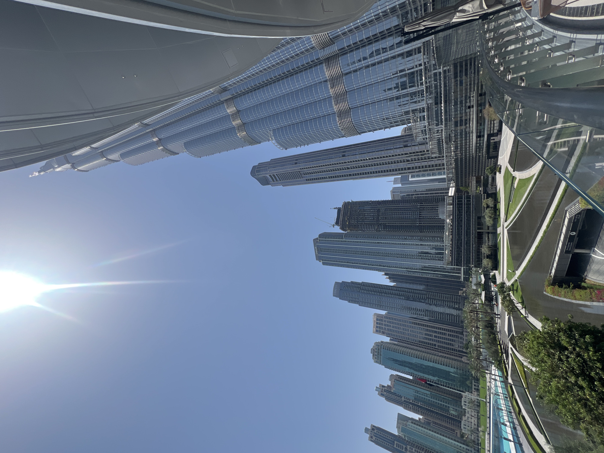 A panoramic cityscape view of Dubai captures the iconic Burj Khalifa towering over a modern urban landscape under a clear, bright blue sky. The image is taken from an elevated perspective, possibly a walkway or another building, looking towards the impressive skyline.
In the left foreground, a sleek, gray architectural structure with horizontal lines frames the top of the image. Below it, the distinctive, multi-tiered, and spiraling design of the Burj Khalifa dominates the left side, its reflective glass and steel facade gleaming in the sunlight. Numerous other skyscrapers, both completed and some still under construction with visible cranes, line a curving waterway or canal that snakes through the urban environment on the right.
The bright sun, positioned in the upper left of the frame, casts a strong light and creates a noticeable lens flare. Along the banks of the waterway, well-maintained greenery, including palm trees and manicured lawns, provides a natural contrast to the towering concrete and glass structures. Pathways and roads are visible alongside the water. A modern glass barrier or railing is present in the immediate foreground on the right, suggesting the vantage point.
Visible text includes "EMAAR" on the side of at least two buildings in the distance, identifying the developer, and a "P" for parking on a sign near the canal. The scene depicts a vibrant, modern city in broad daylight, with no people visible, emphasizing the architectural grandeur and urban planning of Dubai.