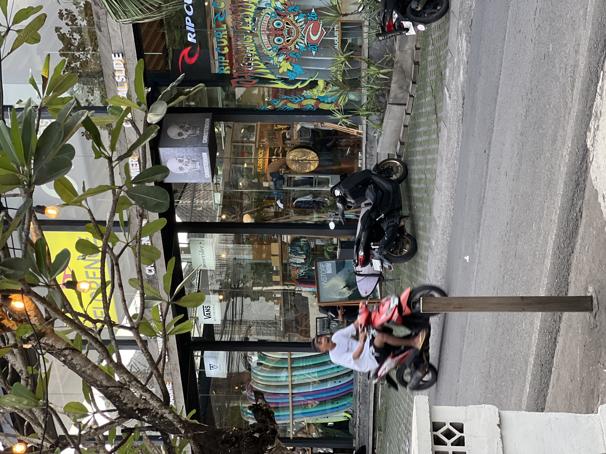This is a street-level, medium-distance shot of a retail storefront in Kuta Selatan, Indonesia. The storefront features a large glass facade displaying various surfboards, clothing racks, and signage for brands like Vans, Christenson, and Rip Curl. A prominent banner on the right displays a colorful graphic with text that appears to be "BALI INDONESIA" and "2026 AUGUST". Two men are visible on scooters on the paved street in front of the store. One man in a white t-shirt and dark shorts rides a red scooter in the foreground, while another figure, partially obscured by foliage, is seated on a black scooter behind him. The street is lined with planters and a wooden post stands near the sidewalk. Trees with green leaves partially obscure the view of the building and sky. The lighting suggests it is daytime, possibly late afternoon, with warm tones from illuminated bulbs hanging from the tree.