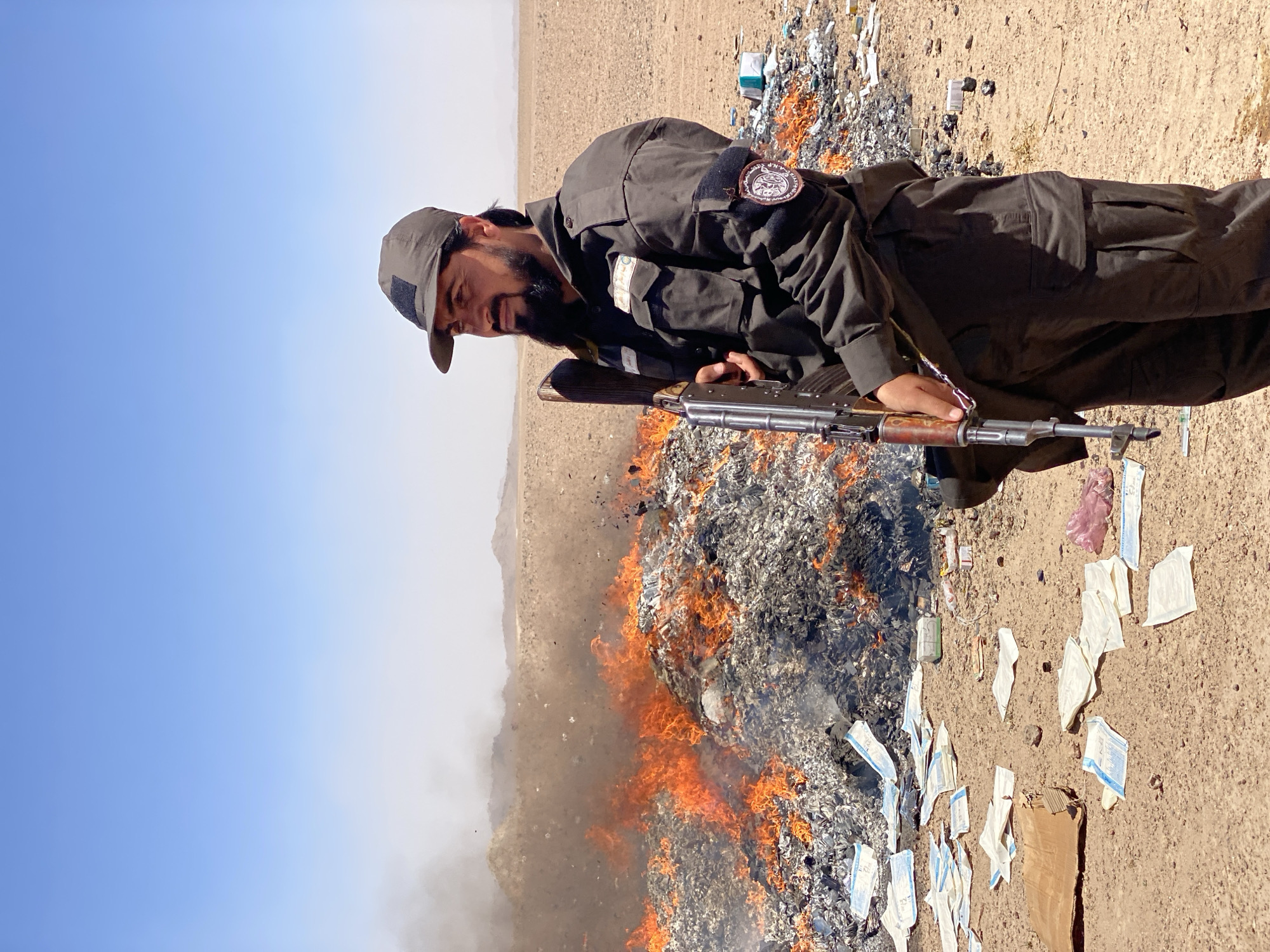 In Mandisar, Afghanistan, under a clear blue sky and a hazy horizon, a man stands in a desert landscape. He is dressed in a dark uniform with patches on the sleeves and wears a dark cap. He holds a rifle with a wooden stock and metal barrel. To his left, a large pile of what appears to be refuse is burning, with flames and smoke rising into the air. Scattered around the burning pile on the sandy ground are various items, including white rectangular objects with blue markings, a piece of brown cardboard, and a pink plastic bag. The overall environment suggests an arid, open area.