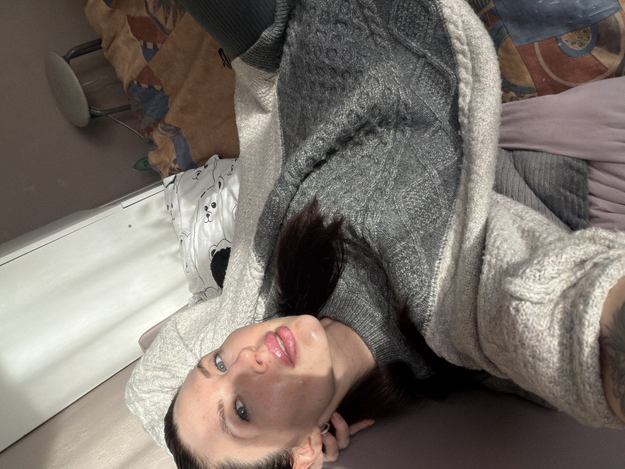 A medium close-up, high-angle shot captures a young adult woman lying on her back in an indoor setting, likely a bedroom, in Perm, Russia. She is looking directly at the camera. The woman has fair skin, dark hair, and striking blue-green eyes. Her lips are slightly parted and appear glossy. She is dressed in a dark grey knitted sweater with a cable-knit pattern, layered under a lighter grey or off-white knitted cardigan or hoodie that features a ribbed texture and cable knit details around the edge. A tattoo is visible on her left arm.
The background shows various domestic elements. To the left, a white pillow with cartoonish black bear faces is visible on the bed, next to a plain light-colored surface, possibly a shelf or desk. To the right, a bed is covered with a patterned duvet featuring geometric shapes in brown, blue, and orange tones, and a solid light purple/mauve sheet or blanket. A light-colored stool with metal legs is partially visible in the upper left background.
Strong natural light from an unseen window casts distinct shadows and bright highlights on the woman's face and the surrounding surfaces, indicating it is daytime. No specific location cues for Perm, Russia are visible within the image itself beyond the general domestic environment. There is no visible text in the image.
