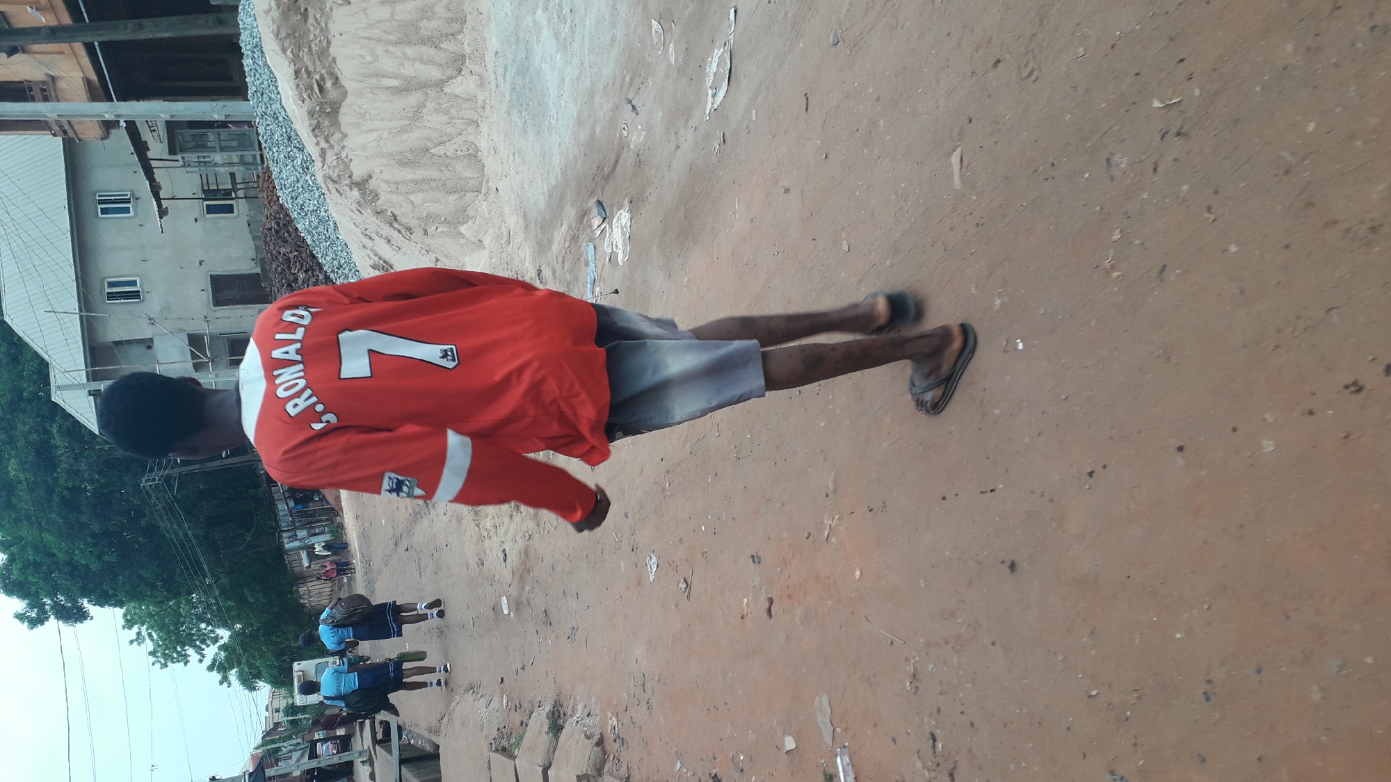 A young man wearing an orange soccer jersey with the name "S. RONALD" and the number 7 on the back stands on a dirt road in Obosi, Nigeria. He is also wearing grey shorts and sandals. In the background, there are buildings, trees, and other people walking. A large pile of construction materials is visible in the background, next to the building. The road is unpaved and appears to be under construction or renovation.
