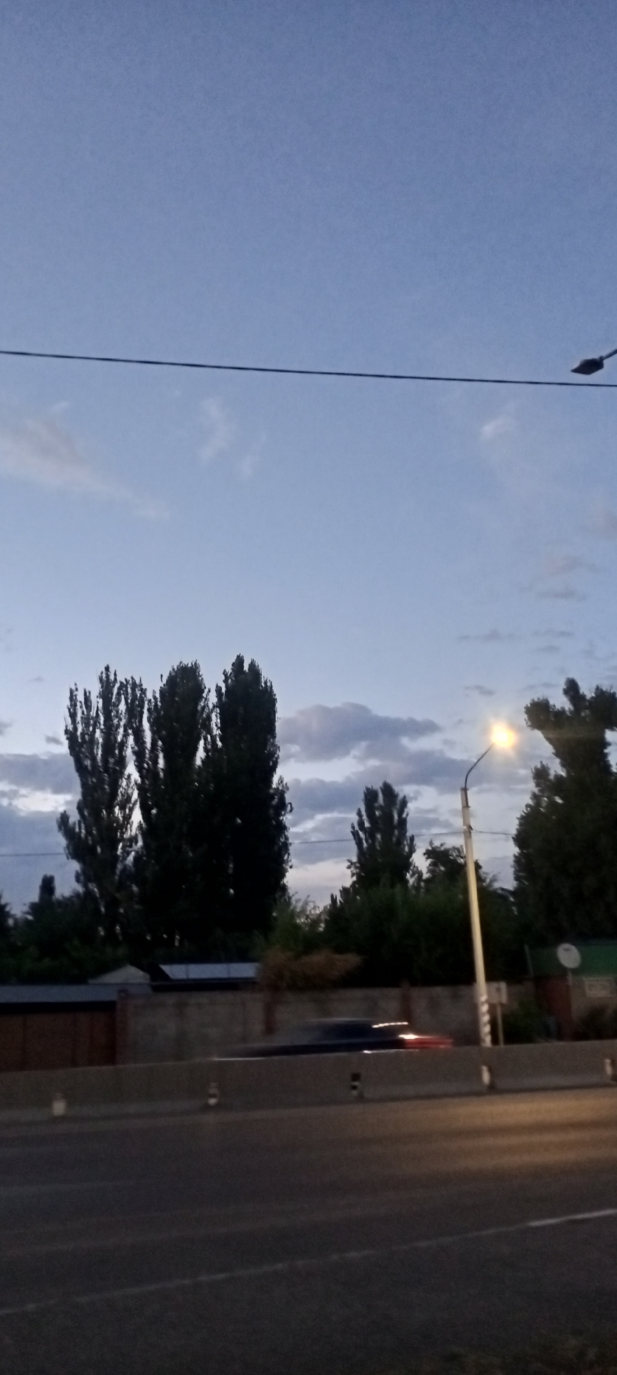 The image captures a street scene in Romanovka, Kyrgyzstan, likely during twilight or early evening. A street lamp is illuminated on the right side of the frame, casting a warm glow. In the background, several tall, slender trees stand silhouetted against a pale blue sky with scattered wispy clouds. A low concrete barrier divides the road, and a dark-colored vehicle is blurred due to motion on the road, with its taillights visible. The overall atmosphere suggests a quiet, suburban or semi-urban environment.