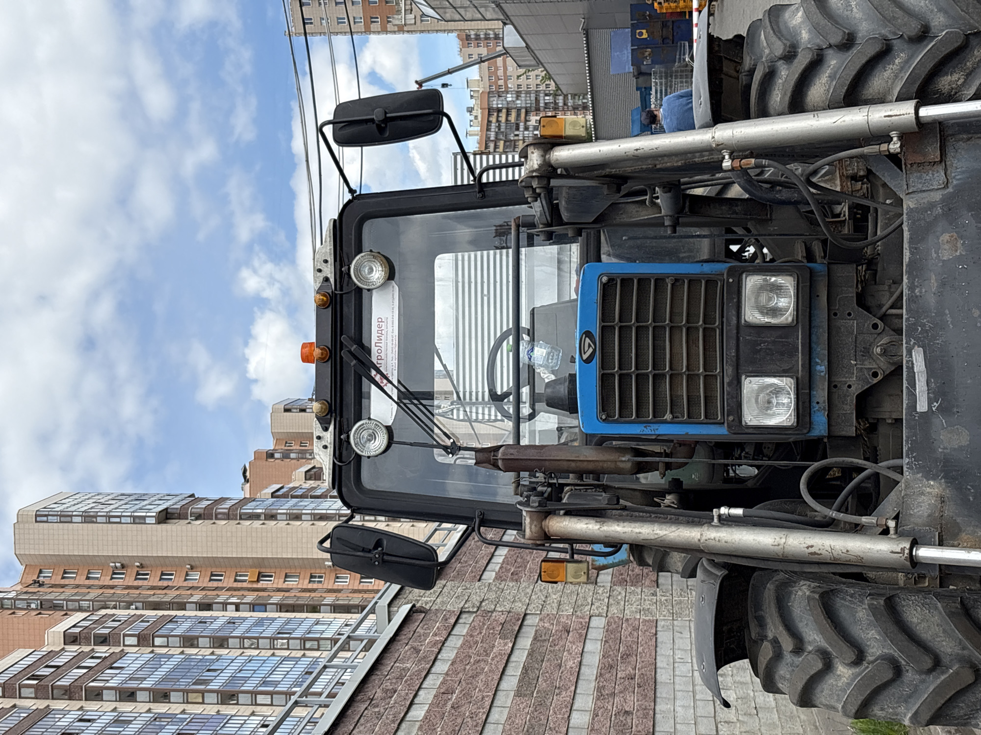 An industrial vehicle, resembling a tractor or specialized utility machine, is prominently featured in an urban setting. The vehicle is largely blue and black, with a robust design including large, deeply-treaded tires, a protective grille on the front, and two rectangular headlights. A cabin with a large windshield, equipped with side mirrors and orange indicator lights, is visible. Hydraulic cylinders and hoses are detailed near the front axle.
The vehicle appears stationary, possibly parked on a paved surface with patterned grey and reddish-brown stones. In the background, multiple multi-story buildings rise against a partly cloudy sky. One building has a distinctive stepped or terraced architecture with numerous windows, while another is a more conventional residential or commercial structure. A single person, dressed in a blue shirt, is partially visible in the mid-ground behind the vehicle, near some industrial equipment or materials.
Notable details include text on a sticker inside the vehicle's cab, which reads "АГРОЛИДЕР" (AGROLIDER) at the top, followed by smaller text like "ПРОДАЖА, СЕРВИС, ЗАПЧАСТИ" (SALES, SERVICE, SPARE PARTS) and contact information. The scene takes place during daytime under a sky with visible white clouds and blue patches. The overall impression is that of a heavy-duty vehicle parked in a city environment, specifically Saint Petersburg, Russia, based on the provided context.