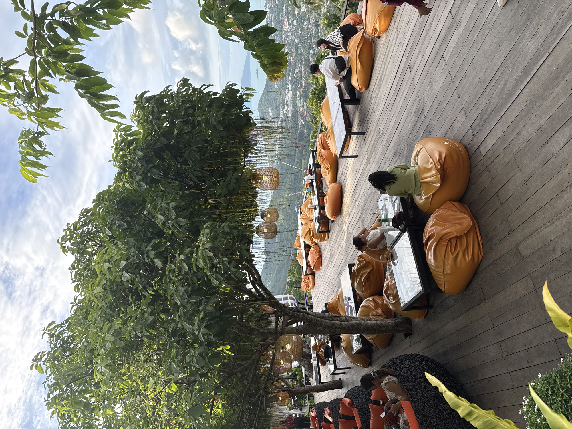 The image depicts an outdoor seating area with a panoramic view of a city and distant islands, likely in the Ko Samui District of Thailand. The foreground features a wooden deck furnished with numerous large, orange beanbag chairs and several low tables. A large tree with lush green foliage, adorned with hanging lights, provides shade over a portion of the seating area. In the midground, several people are seated on the beanbag chairs, some engaged in conversation, while others appear to be dining or relaxing. The background showcases a sprawling cityscape nestled amongst rolling green hills, leading to a hazy ocean horizon with islands visible in the distance. The sky is partly cloudy, with scattered white clouds against a pale blue backdrop. The overall atmosphere suggests a relaxed dining or lounge environment with a scenic overlook.