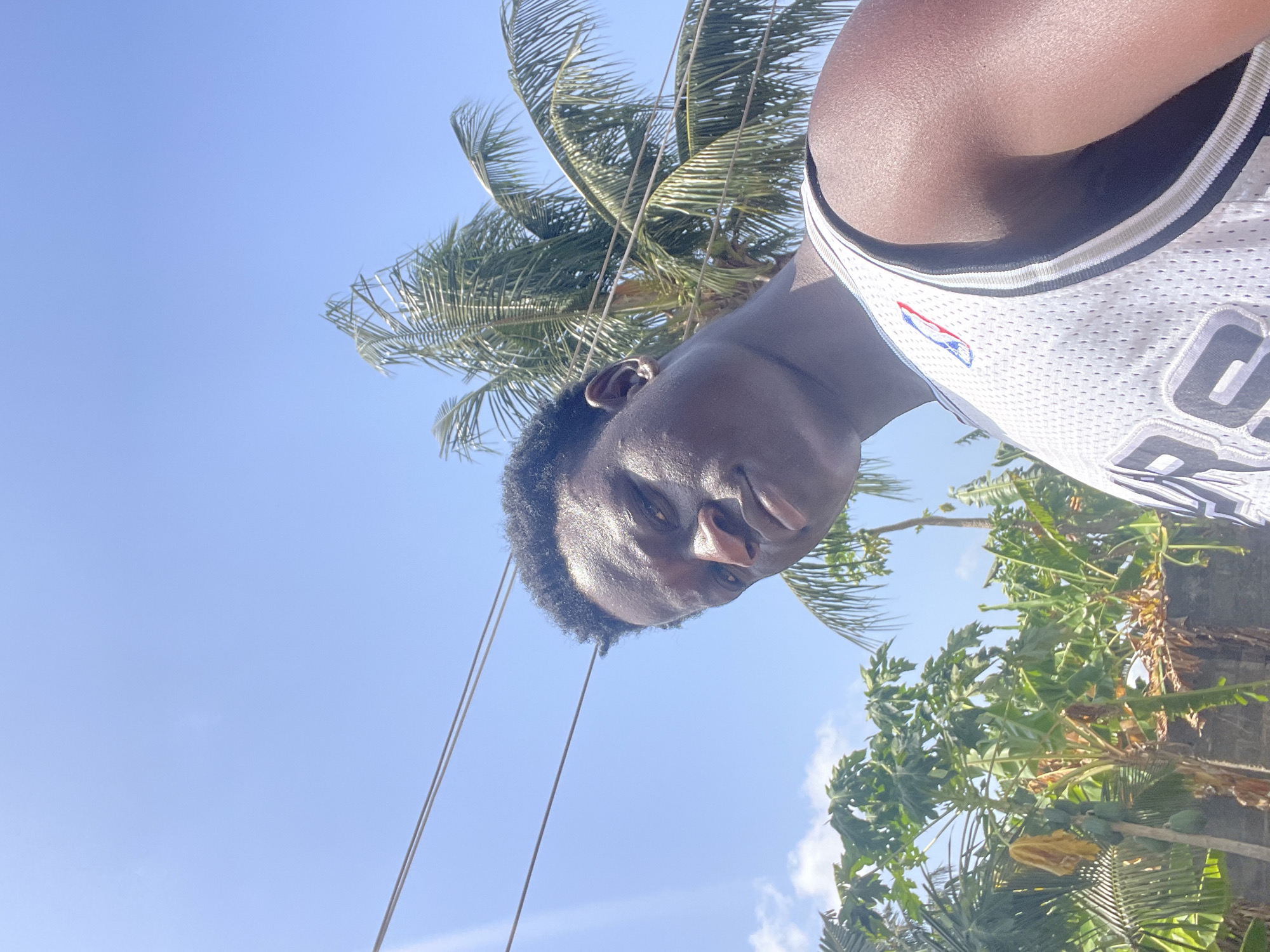 A man is wearing a San Antonio Spurs jersey. He is in Badagry, Nigeria, with a palm tree and other tropical foliage behind him, under a clear blue sky. There are power lines visible.