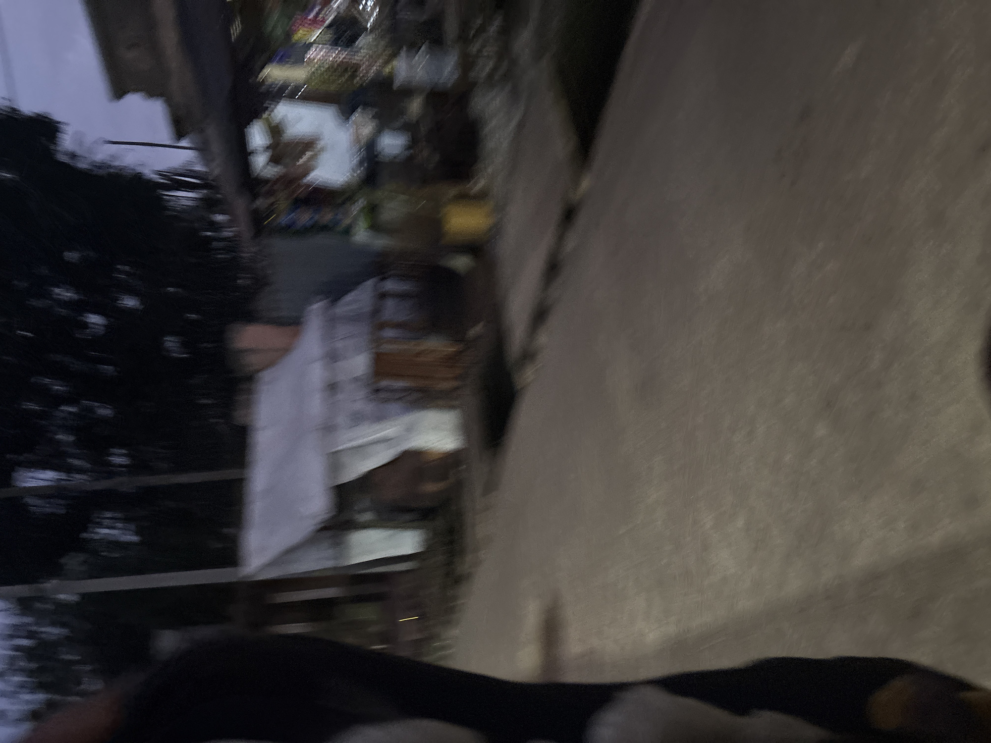 The image captures a blurred street scene in Onitsha, Nigeria, likely taken from a moving vehicle. In the foreground, a dark, out-of-focus object, possibly a person's clothing or a vehicle part, is visible on the left. The middle ground shows a collection of structures that appear to be market stalls or temporary dwellings. These structures have roofs made of corrugated metal or tarpaulin, and some have visible wooden supports. Various objects, including what might be merchandise or supplies, are vaguely discernible within and around these structures. The ground surface is paved with an uneven texture, suggesting a road or marketplace. The background is dominated by dark, indistinct trees and a dimly lit sky, indicating it is either dusk or dawn, or a heavily overcast day. The overall impression is one of a busy, possibly informal, commercial area in Onitsha.