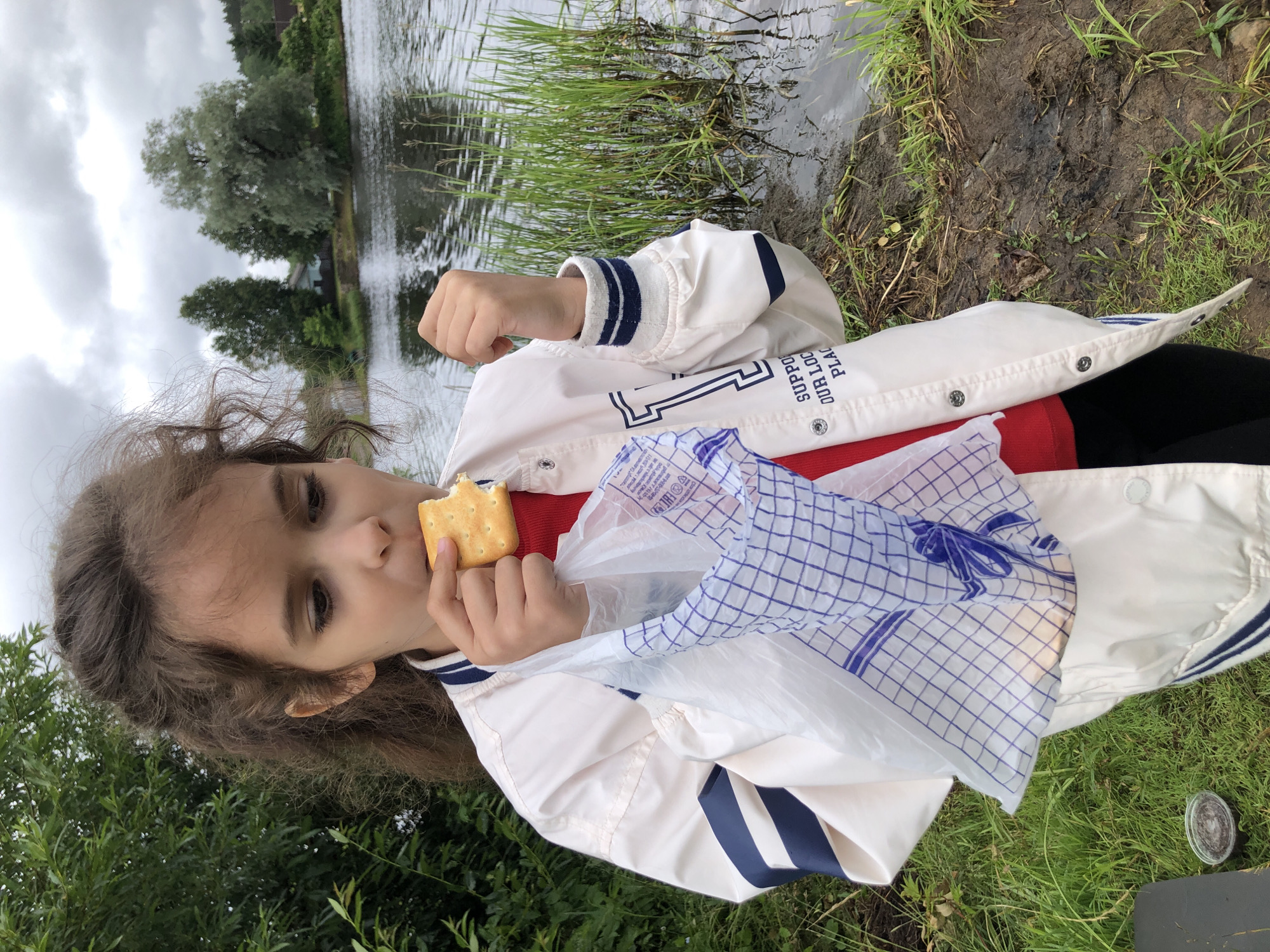 A young girl is captured in a close-up, outdoor shot, eating a cracker. She is wearing a white jacket with navy blue stripes on the sleeves and collar, over a red shirt. In her right hand, she holds a white plastic bag with a blue checkered pattern, and in her mouth, she has a square cracker. Her hair is dark and curly, framing her face as she looks towards the camera. The background suggests a natural setting, with a body of water to the left and lush green grass and trees surrounding her. The water has reflections of the sky and trees, and the edge of the water is lined with tall green grass. The ground is muddy and covered in grass, indicating a damp environment. The sky appears overcast, suggesting a cloudy or possibly rainy day. The image is oriented sideways. Visible text on the plastic bag includes Cyrillic characters and what appears to be a logo or symbol.