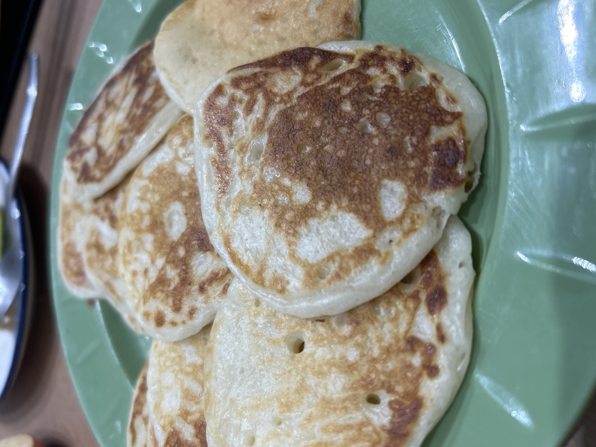 The image displays a close-up, slightly overhead shot of several round, cooked pancakes or fritters arranged on a light green, possibly plastic or melamine, plate. The pancakes vary in size and are light golden-brown with darker, irregularly browned patches, indicating they were pan-fried or griddled. Many of them show small air pockets or holes on their surfaces. The light source illuminates the top surface of the pancakes, creating some glare on the plate's rim. In the blurred background on the left, a sliver of a metallic object, possibly a fork or spoon, can be seen, suggesting an indoor dining or kitchen environment. The scene depicts food prepared for a meal, likely breakfast or a snack. No people or text are visible. The provided context indicates the location is Chaikovsky, Russia.