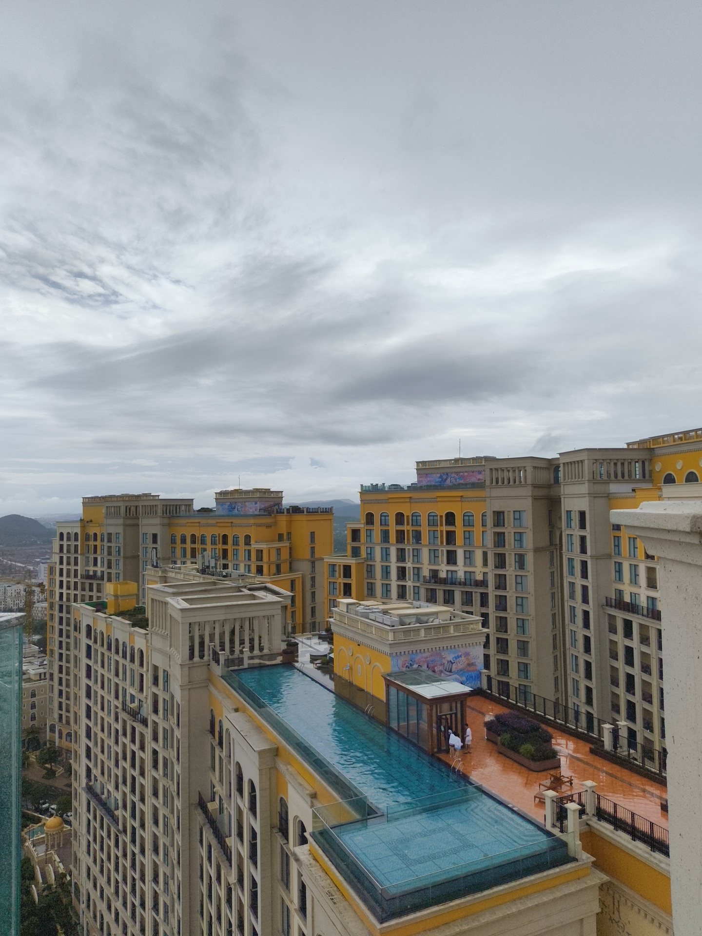 The image shows a collection of tall, modern buildings in a city, likely Phú Quốc, Vietnam, with a cloudy sky. The buildings are predominantly yellow and beige, with numerous windows. One building features a prominent swimming pool on its roof, with a glass enclosure. A few people, dressed in white, are visible near the pool. The architecture has a classical influence, with decorative cornices and archways. The surrounding area appears to be a mix of other buildings and some greenery, with hills visible in the distance. The overall impression is of a resort or residential complex.