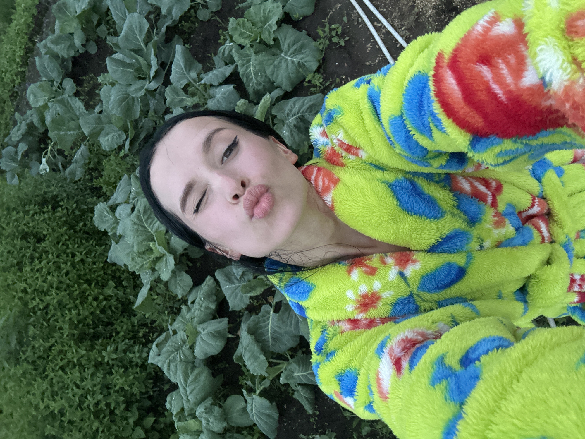 A young woman with dark hair and light skin lies on the ground amidst green foliage, possibly kale or a similar leafy vegetable. She is wearing a bright green, fluffy robe with a colorful tie-dye pattern featuring red, blue, and white accents. Her face is turned towards the camera, with her left eye winking and her lips puckered in a kiss. The lighting suggests it might be daytime, possibly overcast given the soft light on the leaves. The background is filled with the dense greenery of the plants and dark soil. White strings or cords are visible in the upper right corner of the image, possibly part of a garden structure or netting. The scene appears to be in a garden or farm setting. The woman's expression conveys playfulness or affection. There is no visible text in the image. The location is identified as Sosnovo, Russia.
