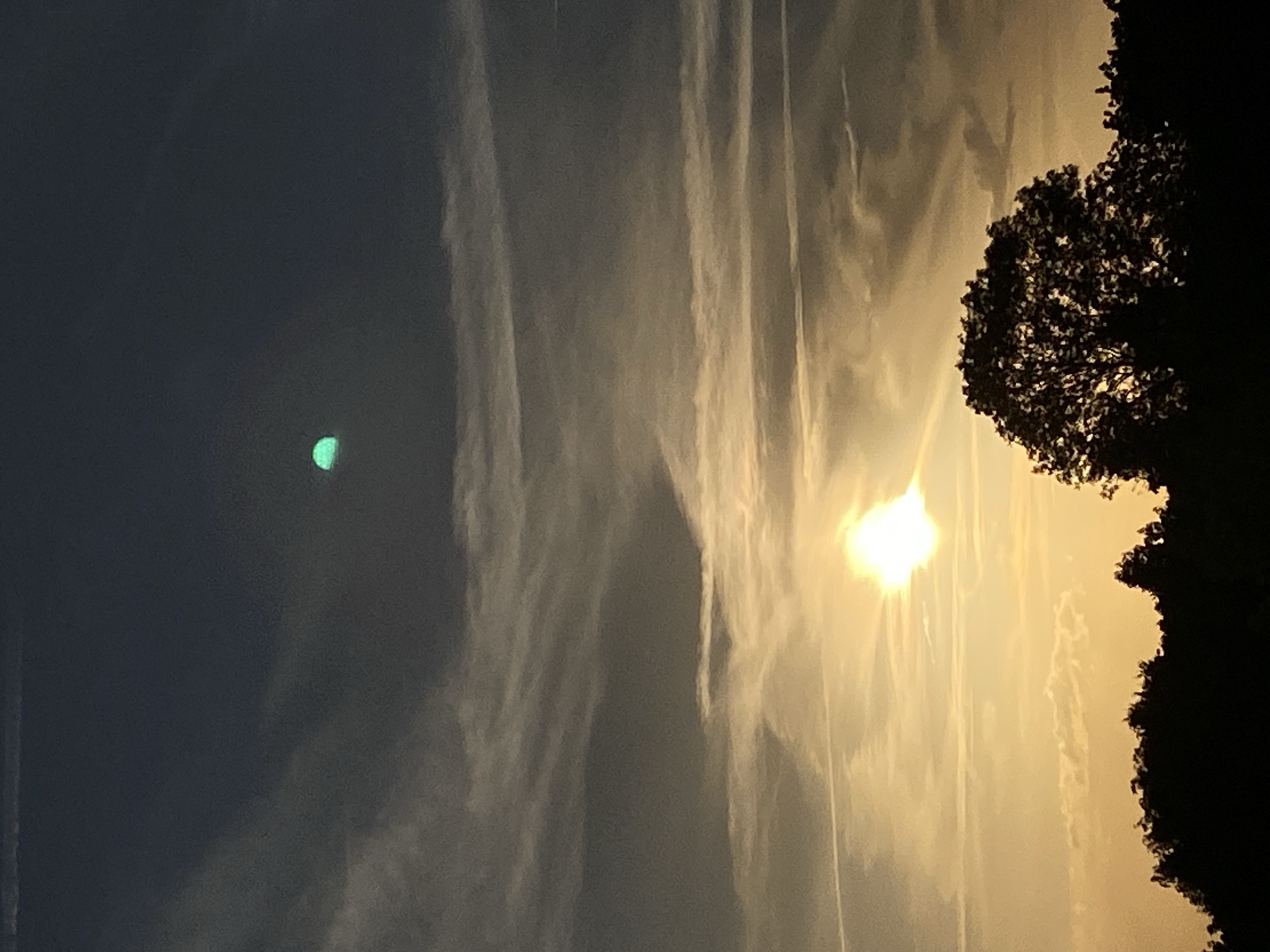 An image of the sky at sunset over Rinteln, Germany, features a sun setting on the horizon, casting a warm, golden light. Silhouetted against this light are the tops of trees and a hill. A notable element in the upper portion of the image is a bright, crescent-shaped object with a distinct green hue. The sky is a blend of dark blue and gray clouds, with streaks of white and orange from the sunset. There is a contrail visible in the upper left corner, suggesting the presence of an aircraft.