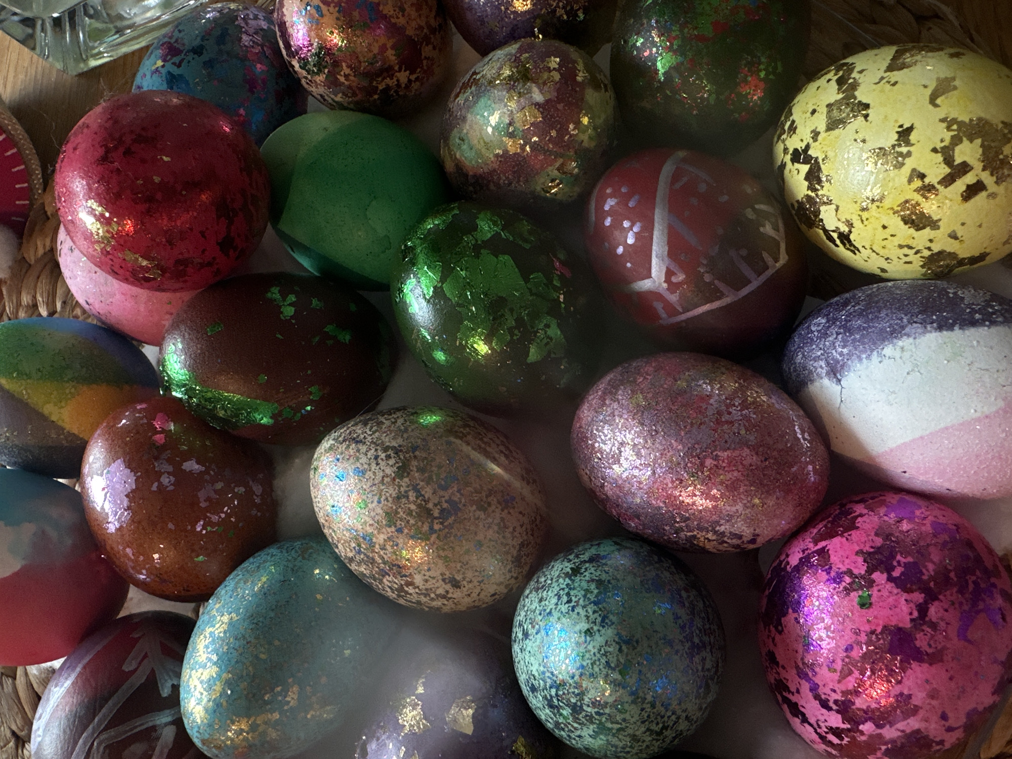 The image shows a close-up of a collection of decorated Easter eggs. The eggs are various colors, including red, green, yellow, brown, blue, purple, and pink. Many of the eggs are decorated with glitter, gold leaf, or paint. The eggs are arranged in a pile. There are no people visible in the image. The location and city are not discernible from the image.