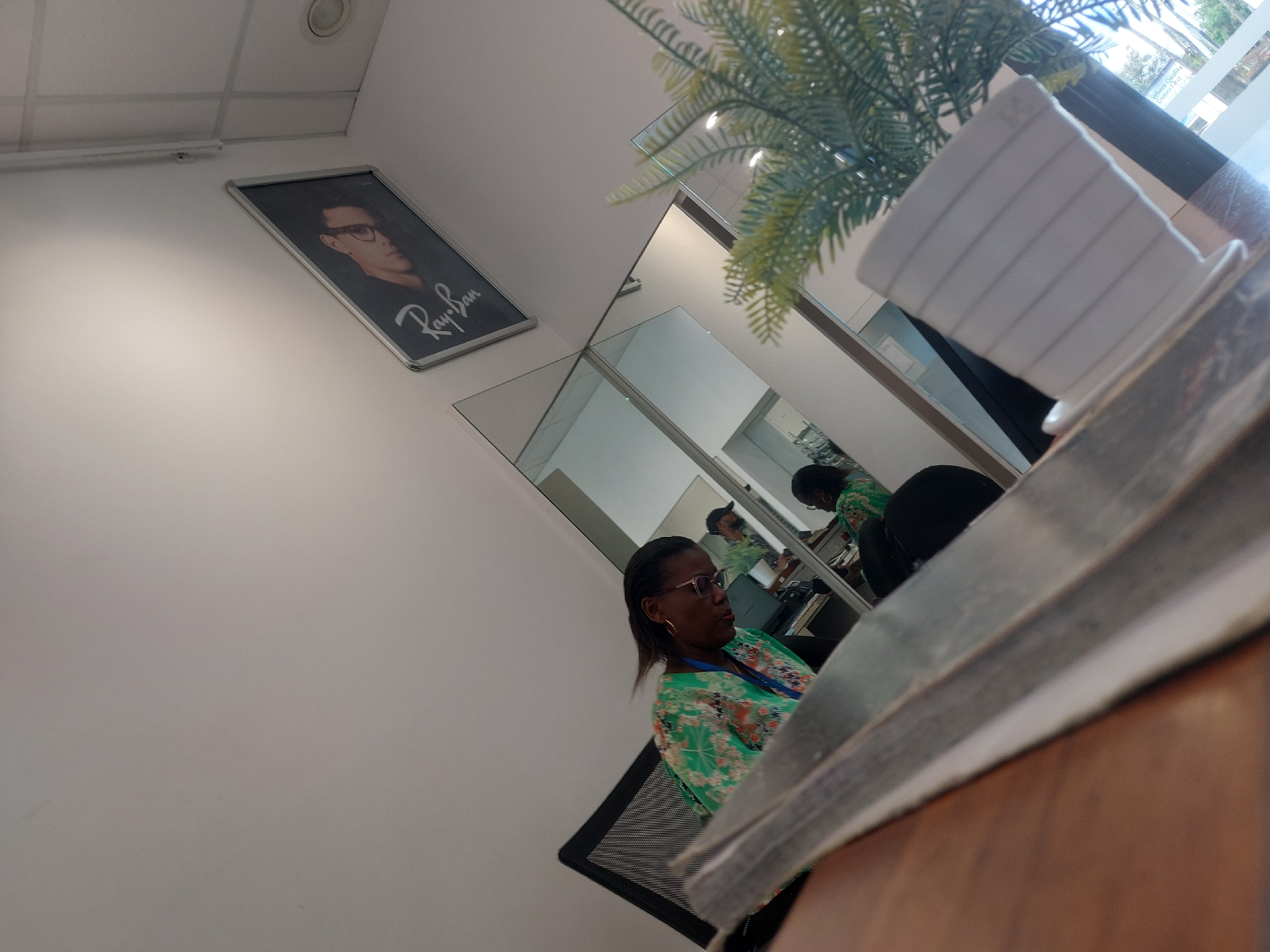 An indoor, diagonally-oriented photograph captures a commercial or office environment, likely a waiting area or reception, with a focus on a seated woman and surrounding decor. The walls are light-colored, possibly white, and a ceiling light fixture is visible at the top left.
In the mid-ground, a dark-skinned woman with glasses and dark hair, wearing a vibrant green and white floral top, sits in a black office chair. Her gaze is directed towards her right. Behind her, multiple large mirrors reflect parts of the room, including her own reflection and what appears to be a workspace with a computer, possibly staffed by another person or another angle of her reflection.
To the upper left, a framed portrait of a man wearing glasses is mounted on the wall. Below the man's image, the brand "Ray-Ban" is prominently displayed in white script. In the upper right of the image, an artificial green plant with palm-like fronds partially obstructs the view. The foreground features a blurry wooden surface, likely a desk or table, on which a white, ribbed cup or container is placed.
The lighting suggests it is daytime indoors. No specific outdoor weather conditions are discernible. The woman's expression is neutral, and the overall scene depicts a typical indoor professional setting in Dar es-Salaam, Tanzania.