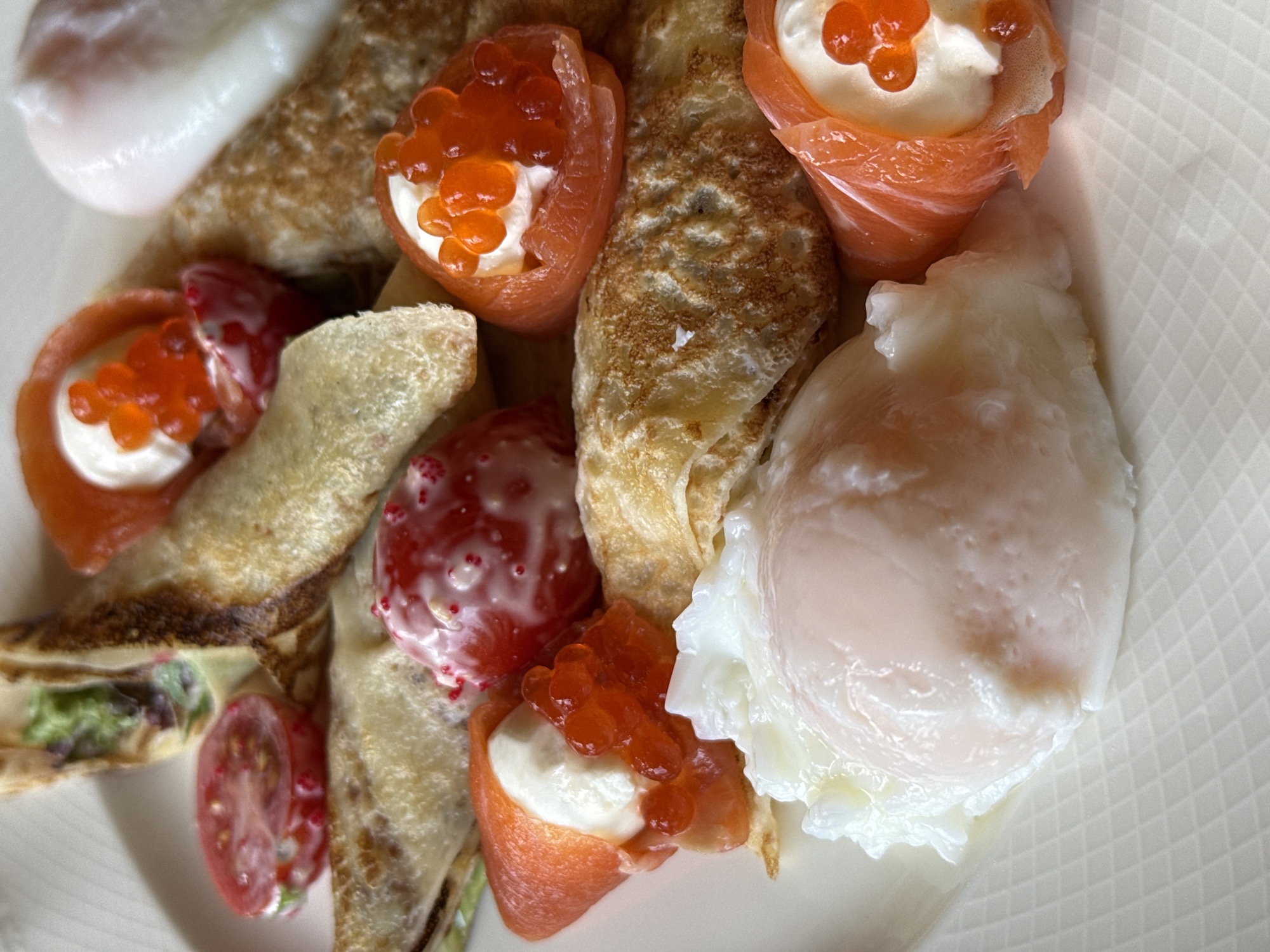 A close-up, overhead shot of a white plate with a portion of a meal. The meal consists of several crepes, some folded, some rolled. Toppings include poached eggs, slices of tomato drizzled with a white sauce and red sprinkles, smoked salmon with cream cheese and red caviar, and what appear to be more tomatoes with cream cheese and red caviar. The lighting is bright, suggesting an indoor setting. The background is a white plate with a subtle grid pattern.