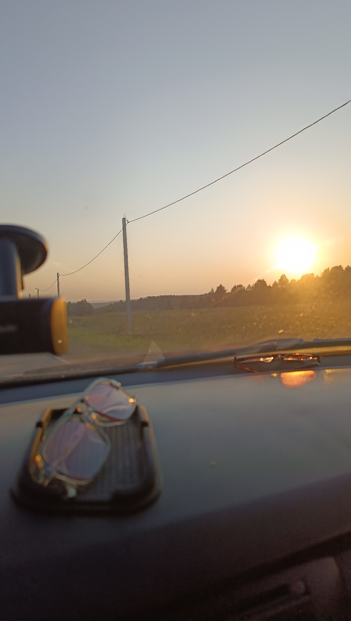 The image captures a sunset viewed from inside a vehicle, likely on a road outside of Chumnaya, Russia. The foreground shows a dashboard with a dashcam mounted on the left side and a pair of sunglasses resting in a holder on the right. The sunglasses are reflecting the warm hues of the setting sun. Through the windshield, a rural landscape is visible, characterized by a field stretching towards a treeline. In the background, utility poles and power lines run parallel to the horizon, with the sun low in the sky, casting a golden glow over the scene. The sky transitions from a pale blue at the top to a warm orange and yellow near the horizon. The overall impression is one of a serene, end-of-day drive through the countryside.