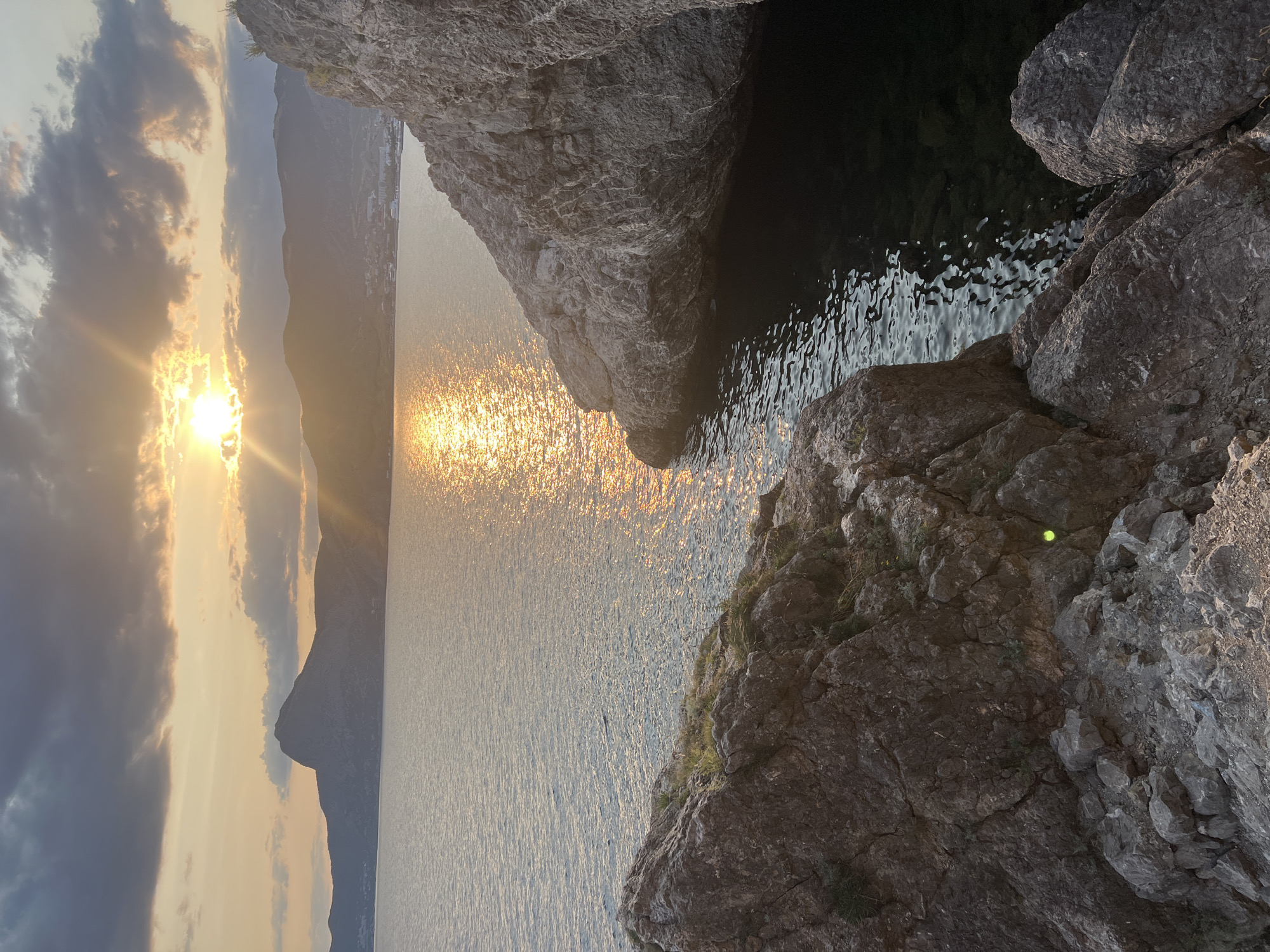 The image depicts a scenic coastal view at sunset near Sudak, Ukraine. The foreground is dominated by rugged, textured rock formations, one on the left and another larger one on the right, creating a natural frame for the scene. These rocks are rough and appear to be weathered, with some sparse vegetation growing on them.
Beyond the rocky foreground, a calm body of water stretches towards the horizon. The water reflects the warm, golden light of the setting sun, creating a shimmering path across its surface. In the middle ground, a range of hills or mountains forms the backdrop. The tallest peak, with a distinct, somewhat rounded summit, is visible on the left side of the image. Along the coastline of these hills, a settlement or town can be discerned, with buildings and lights indicating its presence.
The sky is a mix of dramatic clouds and clear areas. Dark, heavy clouds are present in the upper left quadrant, contrasting with the bright, radiant glow of the sun breaking through the clouds on the right side. Sunbeams emanate from the sun, casting a warm, diffused light across the sky and water. The overall atmosphere is tranquil and picturesque, characteristic of a coastal sunset. No people or animals are visible in the image.