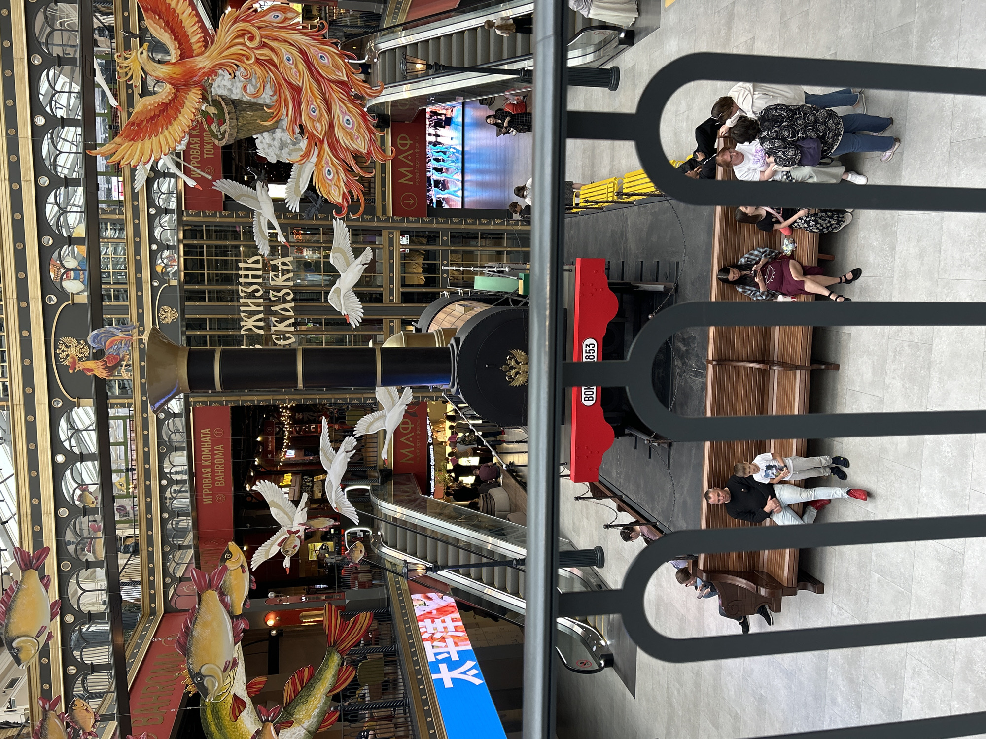 This is a vertical, eye-level, medium shot of a busy indoor marketplace or shopping mall, featuring a decorative display and several people. The primary focus is a large, ornate decoration suspended from the ceiling, depicting a fiery phoenix and several white birds that resemble cranes. Below the phoenix, a train car with "853" visible on its side is present.
In the background, escalators lead to upper levels, with people visible ascending and descending. A large screen displays moving images of a dance performance, with the text "МАФ музей орт и факты" and "музей нового формата. От чудес" displayed prominently. Other text includes "ИГРОВАЯ КОМНАТА BAHROMA", "К КИНОТЕАТР", and "ЖИЗНЬ". Decorative elements like large fish cutouts and rooster figures are also part of the display.
Several groups of people are seated on benches. One group consists of three women and a child, appearing to be in conversation. Another group includes a man and a child sitting together, and further down the bench, two men are seated. The overall atmosphere appears lively and bustling, typical of a public indoor space. The lighting suggests it is daytime.