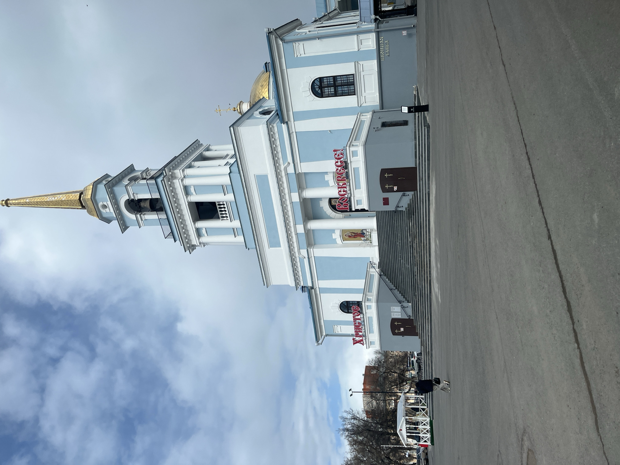 This image captures a large, ornate Orthodox church with a light blue and white facade. The church features multiple white columns, arched windows, and decorative white trim. A prominent golden spire topped with a cross rises from a bell tower on the left, while a smaller golden dome with a cross is visible further back on the main building. Several sets of grey steps lead up to the church's entrances.
The foreground consists of a wide, grey paved area. One person, dressed in dark attire, is walking away from the camera on this paved surface, carrying a light-colored bag. In the background to the right, there is a white tent-like structure, and another individual is faintly visible nearby. Leafless trees line the distant background, suggesting a colder season. The sky is overcast with scattered clouds, indicating daytime.
Visible Cyrillic text on the church facade includes large red letters spelling "ВОСКРЕСЕ!" (VOSKRESE!, meaning "He is Risen!"). Below this, a smaller sign also in red reads "ХРИСТОС" (CHRISTOS, meaning "Christ"). Above a window on the side of the building, the text "ЦЕРКОВНАЯ ЛАВКА" (TSERKOVMYAYA LAVKA, meaning "Church Shop") is visible. The architectural style and text are consistent with the location being Votkinsk, Russia.
