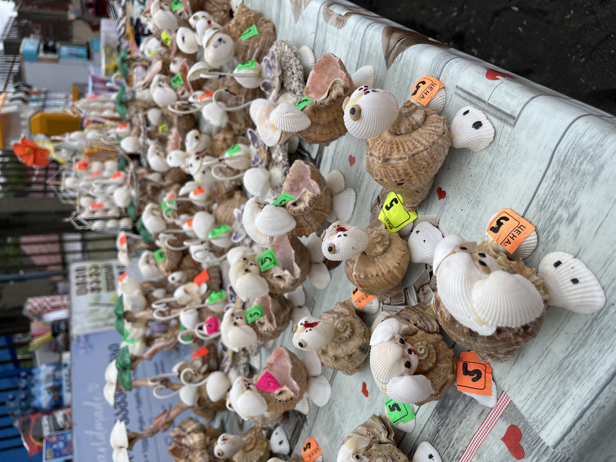 At a street market in Ureki, Georgia, a table displays numerous seashell crafts. The crafts are primarily in the shape of animals, such as turtles and dogs, constructed from various types of shells. Some crafts have small, colorful tags attached, possibly indicating prices. In the background, there are more market stalls and a fence. The lighting suggests it is daytime, and the overall setting appears to be an outdoor commercial area.