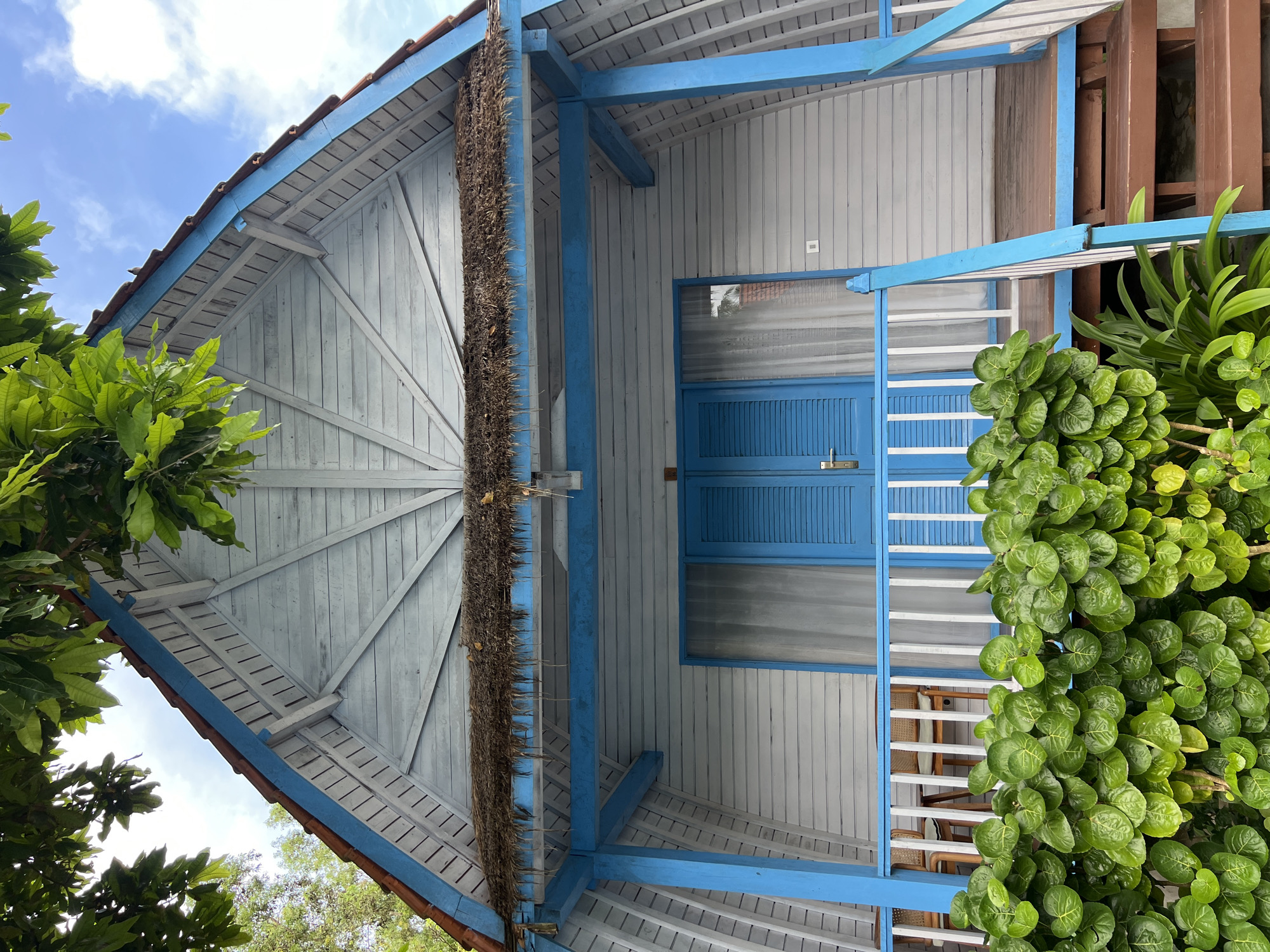 The image depicts the exterior of a building in Nusa Penida, Indonesia, viewed from a low angle. The structure features light gray wooden planks with blue trim on the roof and structural supports. A section of dried vegetation hangs along a vertical support beam. A blue-shuttered window or door is visible in the center. The building has a wooden porch or balcony with white railings. Lush green foliage surrounds the edges of the frame, partially obscuring the building. The sky is visible in the upper left corner.