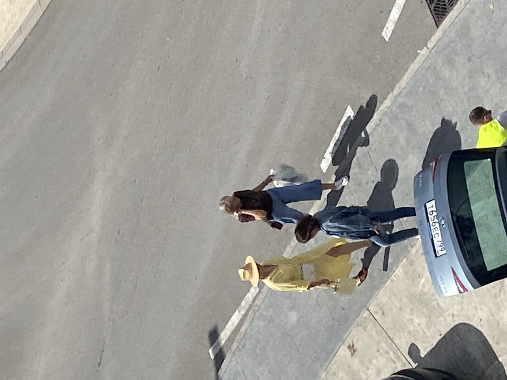 The image presents a high-angle view of a street scene in Budva, Montenegro, under bright sunlight, indicated by the long, distinct shadows cast by people and objects. The paved roadway occupies the left and central parts of the frame, bordered by a sidewalk and curb on the right.
Several individuals are present on or near the sidewalk. In the middle of the frame, a woman wearing a dark top, light blue trousers, and white athletic shoes walks, carrying a light-colored bag. Adjacent to her and slightly ahead, another person is visible in a blue denim jacket, blue trousers, and red shoes. Closer to the parked car, a person in a light yellow top and a straw hat is seen. A fourth person, wearing a bright yellow top, is partially visible near the rear of the car on the far right.
A silver or light blue hatchback car is parked parallel to the curb. Its rear license plate is clearly legible and displays the alphanumeric sequence "T656EC199". White painted lines delineate parts of the road, and a metal storm drain grate is visible on the sidewalk in the upper right quadrant. The individuals appear to be walking, suggesting everyday pedestrian activity in a sunny, urban environment.