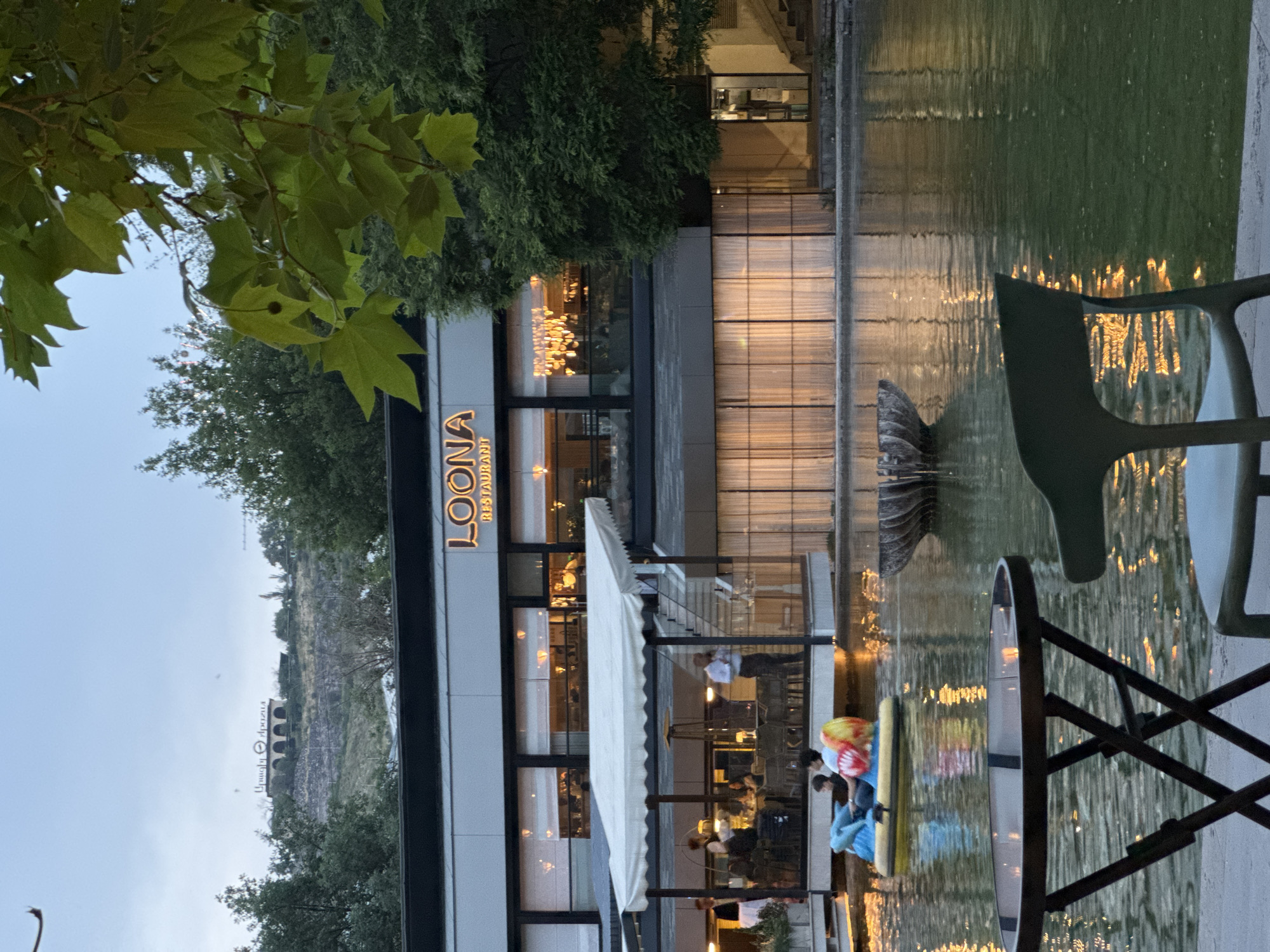 Captured in Yerevan, Armenia, the image features the "LOONA restaurant", its illuminated sign visible. The building’s modern architecture is reflected in a body of water, possibly a pond or artificial lake, with chairs and tables set nearby, suggesting an outdoor dining area. Diners and staff are visible inside the restaurant. The surroundings include trees and, in the background, a historical structure. People are also enjoying the water with floating devices.