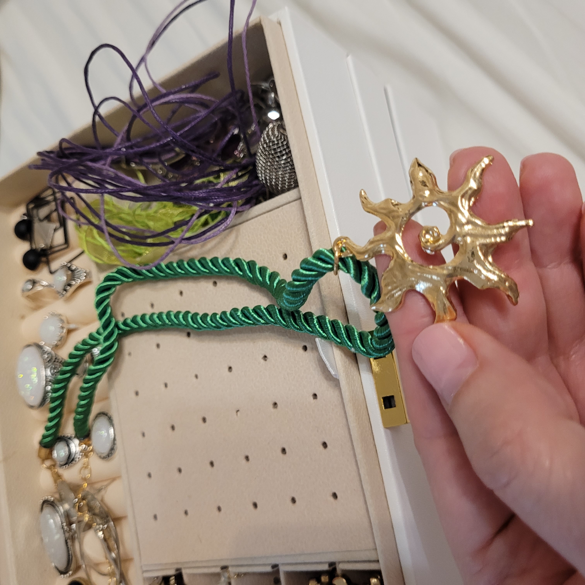 A close-up, high-angle shot captures a person's hand holding a piece of golden jewelry over an open jewelry box. The hand, with fair skin and well-maintained nails, firmly grasps a distinctive gold-colored pendant or brooch. This piece of jewelry is abstract and organic in design, resembling a swirling sun or star with radiating, textured rays. It connects via a small gold loop to a thick, braided green cord that looks like a necklace.
The open jewelry box occupies the left and middle of the frame. It appears to be made of light-colored (possibly beige or cream) material, likely faux leather or velvet, with white dividers. Inside the box, various items are visible. The top left compartment contains a tangled mess of purple and light green cords or threads. A silver-toned, spherical object with a textured, almost mesh-like surface is also partially visible in this section.
The middle section of the jewelry box features padded inserts designed to hold rings and earrings. Several silver-toned rings are visible, each adorned with a large, oval, iridescent white stone, possibly opal or mother-of-pearl. The braided green cord necklace, to which the golden pendant is attached, is draped over these ring holders. The surface of the padded insert has small, regularly spaced holes, likely for holding stud earrings, though no earrings are clearly visible in those specific slots.
The overall scene suggests someone is either selecting jewelry from their collection or organizing it. The lighting is bright and even, indicating an indoor setting. There is no visible text in the image.