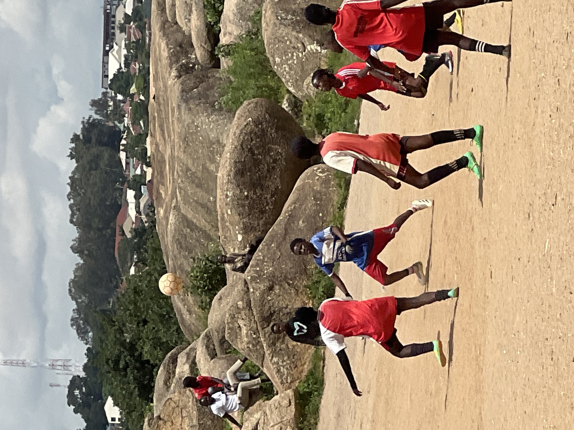 The image depicts an outdoor scene in Jos, Nigeria, featuring a group of young people playing soccer amidst a rocky, uneven terrain with scattered vegetation and distant buildings. Several children, predominantly boys, are actively engaged in a game of soccer, dressed in sportswear. One boy in a red and white jersey is in mid-air, appearing to head a soccer ball. Another boy in a blue and red jersey is positioned defensively. In the background, other individuals are either watching the game or resting on large rocks. The environment is characterized by large, weathered rock formations and a dusty, sandy ground. The presence of a transmission tower and residential buildings in the distance suggests an urban or semi-urban setting within Jos. The sky is partly cloudy, indicating daytime.