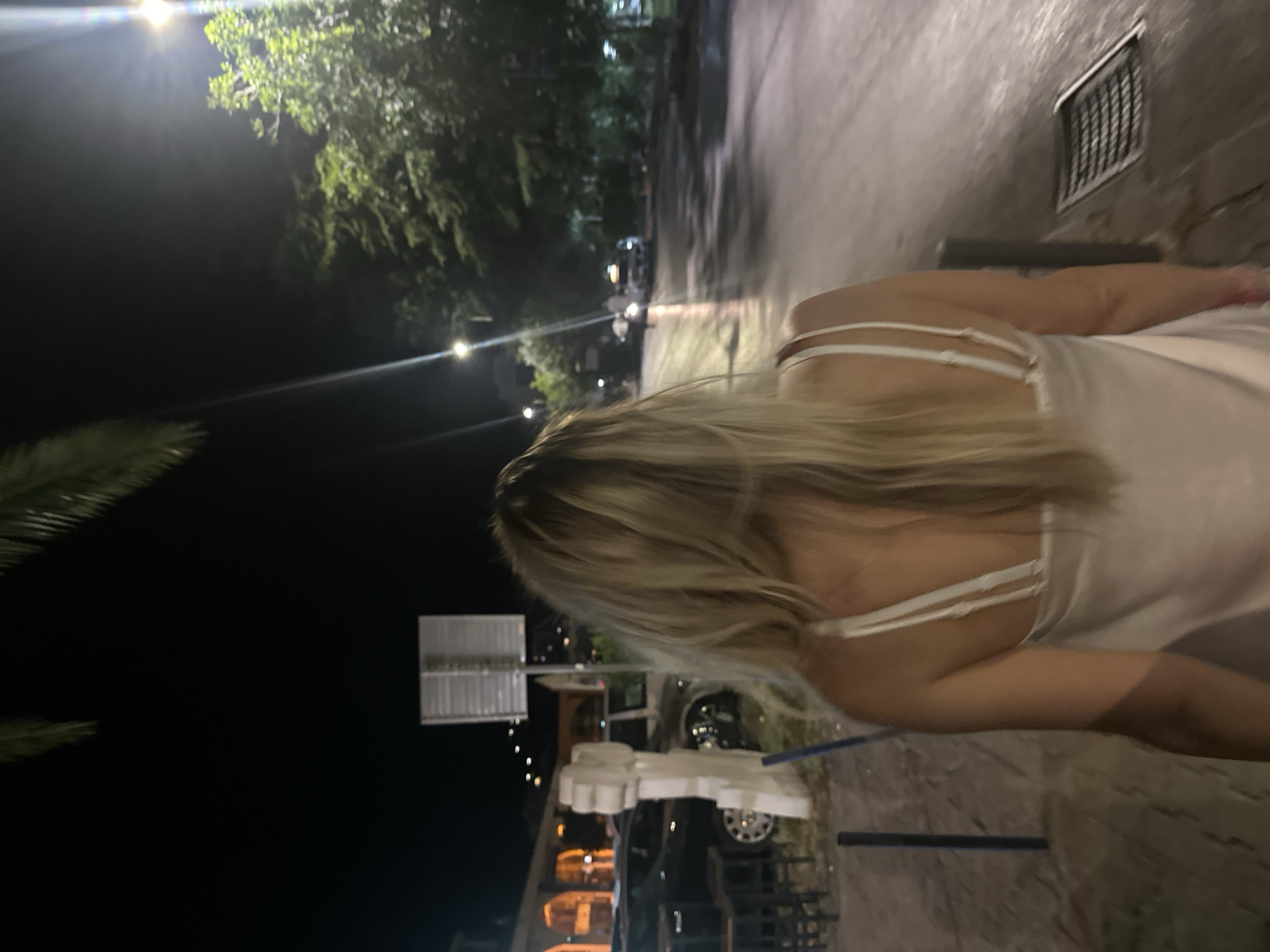 A blonde woman, seen from behind, walks on a wet paved sidewalk at night in Budva, Montenegro. She wears a white slip dress with thin straps. The street is lined with trees and illuminated by streetlights, casting reflections on the wet asphalt. In the background, there are buildings with arched windows, parked cars, and a few people walking further down the street. The overall scene suggests a lively urban environment at night.
