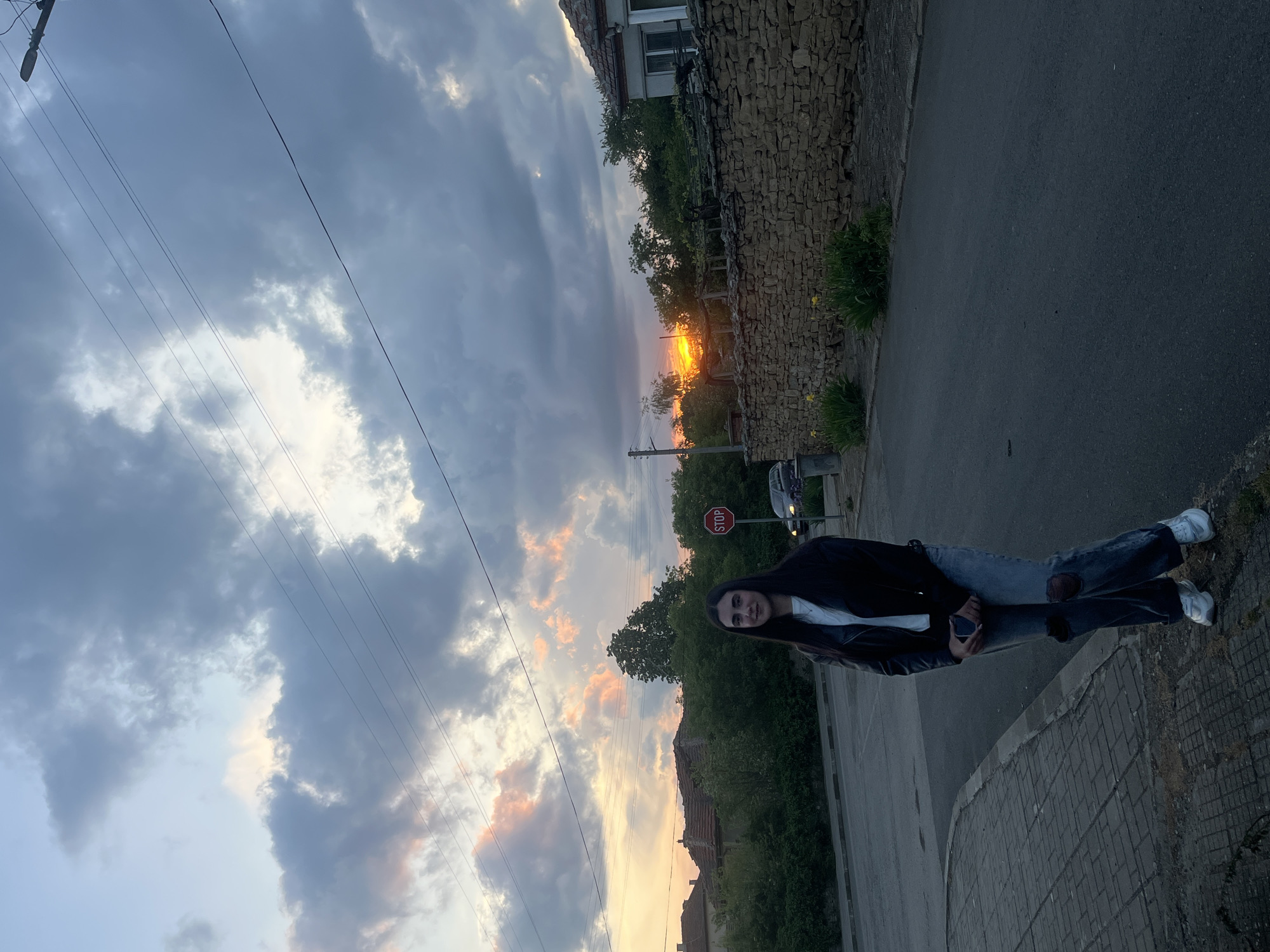 The image shows a young woman standing on a sidewalk in Lozarevo, Bulgaria. She is wearing a black jacket, white shirt, and blue jeans with sneakers. She is holding a phone in her hand. Behind her is a street, and a stone wall with trees and greenery. A "Stop" sign is visible behind the woman. The sky is cloudy with a sunset glow. Buildings are seen in the background. Overhead power lines are also present.