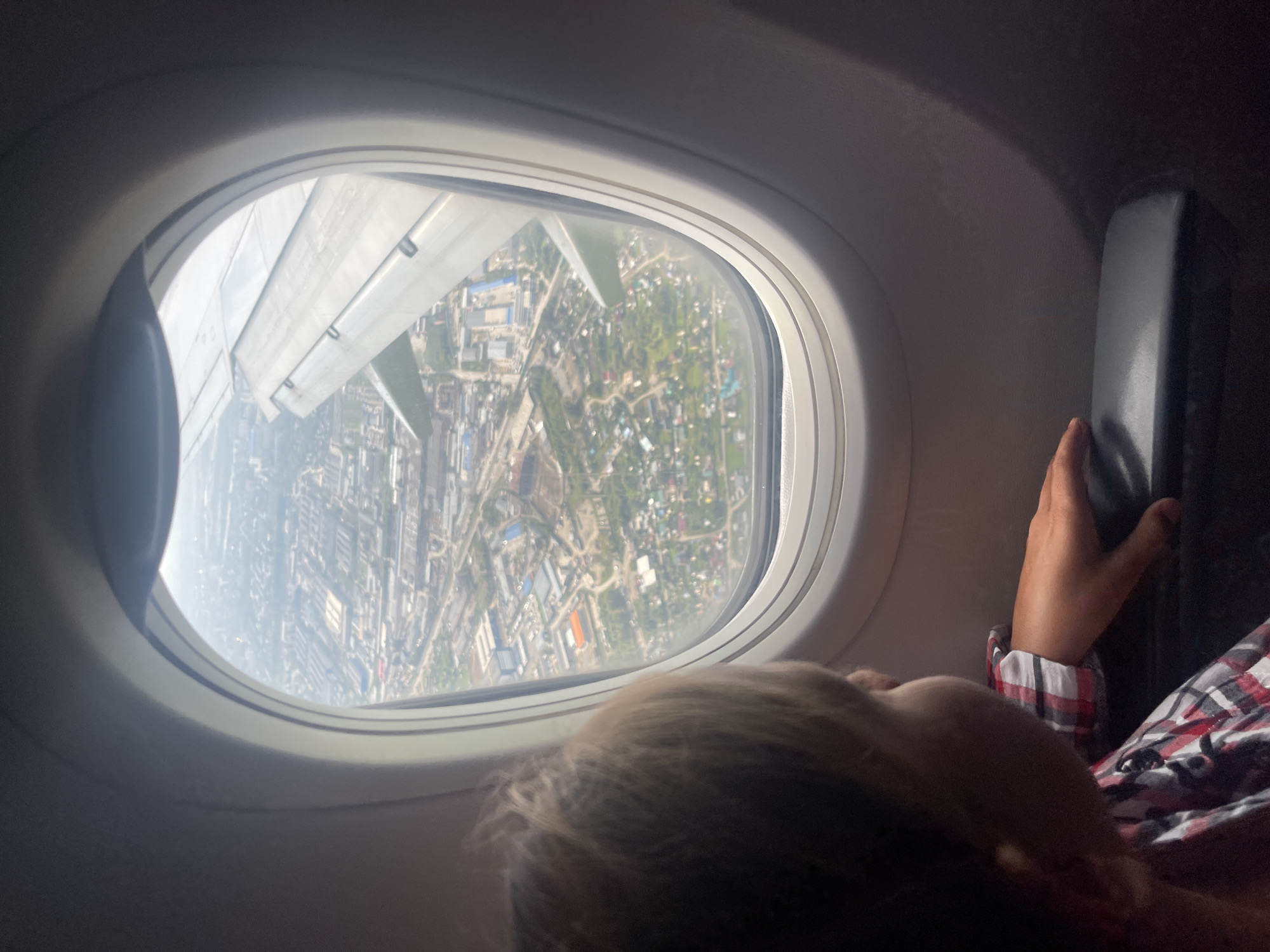 A child's head and shoulder are in the foreground, looking out an airplane window. The child has light brown hair and is wearing a red, white, and black plaid shirt. Their right hand rests on the armrest. Outside the window, a city stretches out below, with buildings, roads, and greenery visible. An airplane wing is also visible, angled upwards. The scene is consistent with being in the air over the city of Sibirskiy, Russia, given the urban and semi-rural landscape visible from above.
