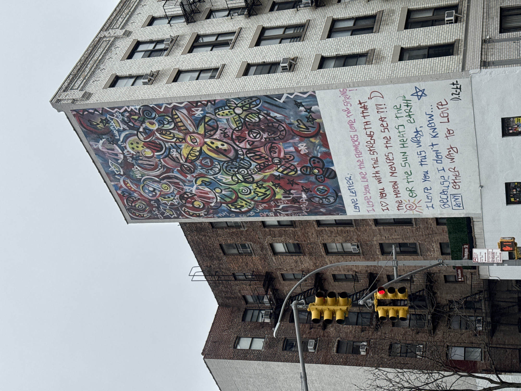 The image shows an urban scene in New York City, United States of America. A large mural, featuring abstract shapes and floral designs in vibrant colors, is painted on the side of a light-colored brick building. To the right of the mural, a white section of the building displays handwritten text. Below the mural, there's a brown brick building. A traffic signal with red and yellow lights hangs between the two buildings. The street sign reads Thompson St and W Houston. The sky is overcast.