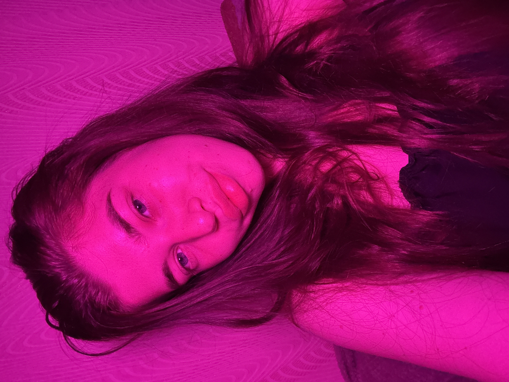 A young woman with long, dark hair is captured in a close-up, selfie-style shot, illuminated by a vibrant pink light. She is looking directly at the camera with a slight smile. Her eyes are a light blue, and her lips are full and slightly parted. She is wearing a black top. The background appears to be a textured wall, also bathed in the pink light. There is no discernible location in the city of Мещёры, Russia visible in the image.
