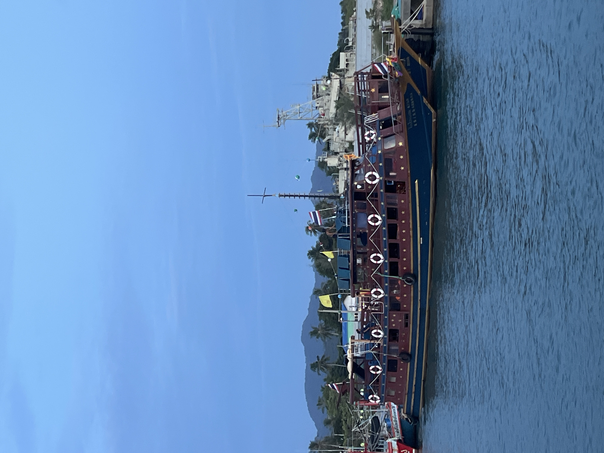 The image shows a harbor scene with two large boats docked side-by-side. The boats are ornate, with dark blue hulls and maroon upper decks decorated with gold trim and star patterns. Several life preservers are visible along the railings of the boats. The boats are moored to a concrete dock on the right side of the image. In the background, there is a hazy silhouette of mountains and palm trees, suggesting a tropical location. A large, complex structure, possibly a lighthouse or industrial facility, is visible further in the background. The sky is a deep blue, indicating either early morning or late evening. The water in the foreground is a deep blue, with gentle ripples on the surface. The overall impression is of a peaceful harbor at dusk or dawn.