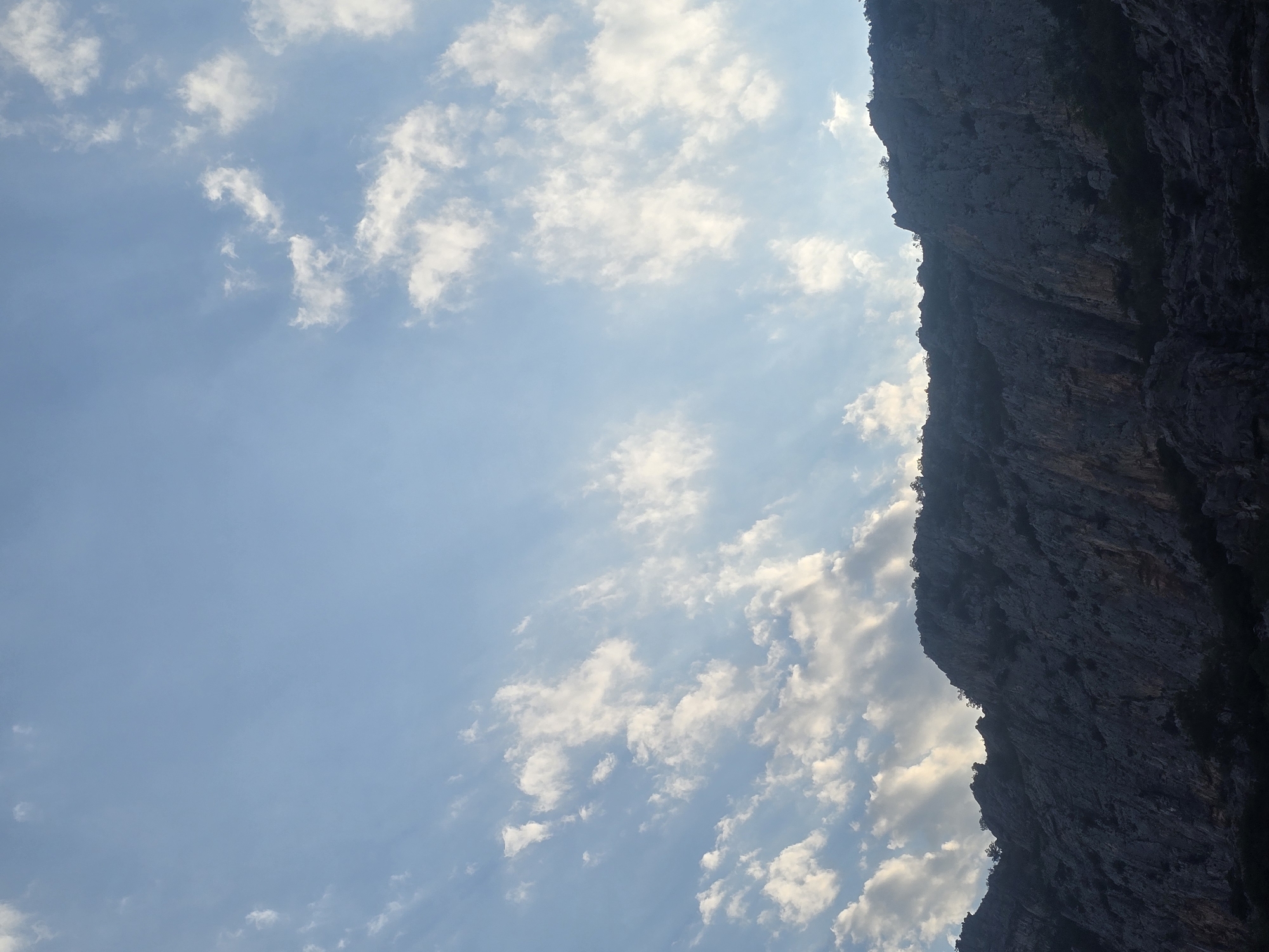 The image displays a rugged, dark mountain range under a vast sky filled with scattered white clouds. The mountain's rocky texture and steep slopes are visible, with sparse vegetation clinging to its upper edges and base. The sky is a gradient of light blue, with the clouds appearing brighter where sunlight catches them. The scene is devoid of any human presence or man-made structures. The photograph appears to be taken from a low angle, looking upwards towards the mountain and sky. The location is stated to be Podgorica, Montenegro, suggesting this is a natural landscape within or near the city.