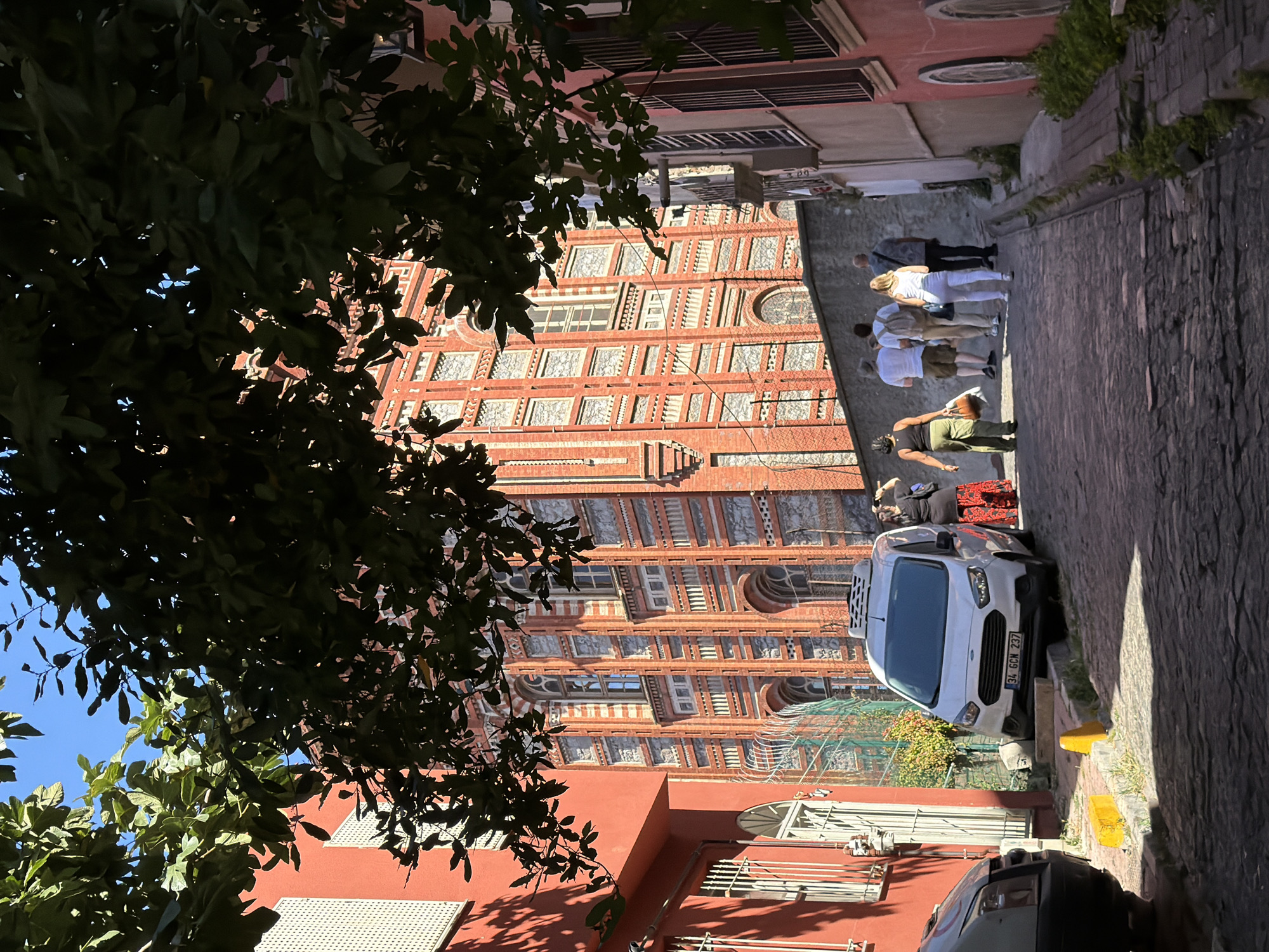 In Istanbul, Turkey, on a cobblestone street lined with historic buildings, a group of tourists walks up a sloping lane. The large, ornate brick building on the right is the Phanar Greek Orthodox College, a prominent landmark in the Fener district. A white van is parked on the left side of the street. People are seen walking in the same direction, with some carrying bags and wearing casual attire. The sun casts shadows on the street, indicating it is daytime. The foliage from the surrounding trees partially obscures the view of the buildings.