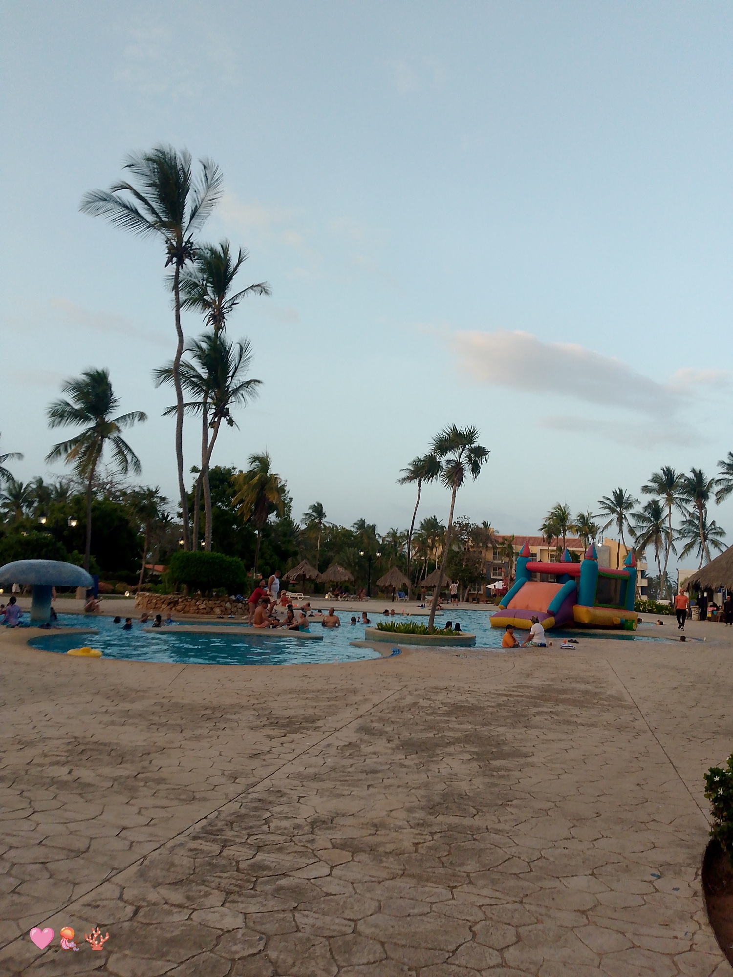 The image depicts a water park scene, likely at a resort, in what appears to be the late afternoon or early evening. The location is speculated to be in Playa El Agua, Venezuela.
The park includes a large irregular-shaped swimming pool filled with people. Many people are swimming while others are sitting at the pool’s edge. To the right of the pool is an inflatable water slide, suggesting a family-friendly environment.
The surrounding area is paved with large patterned stones and includes palm trees that are scattered around the park. Several structures with thatched roofs are also present. The sky is pale blue with light clouds. The overall environment suggests a tropical vacation destination.