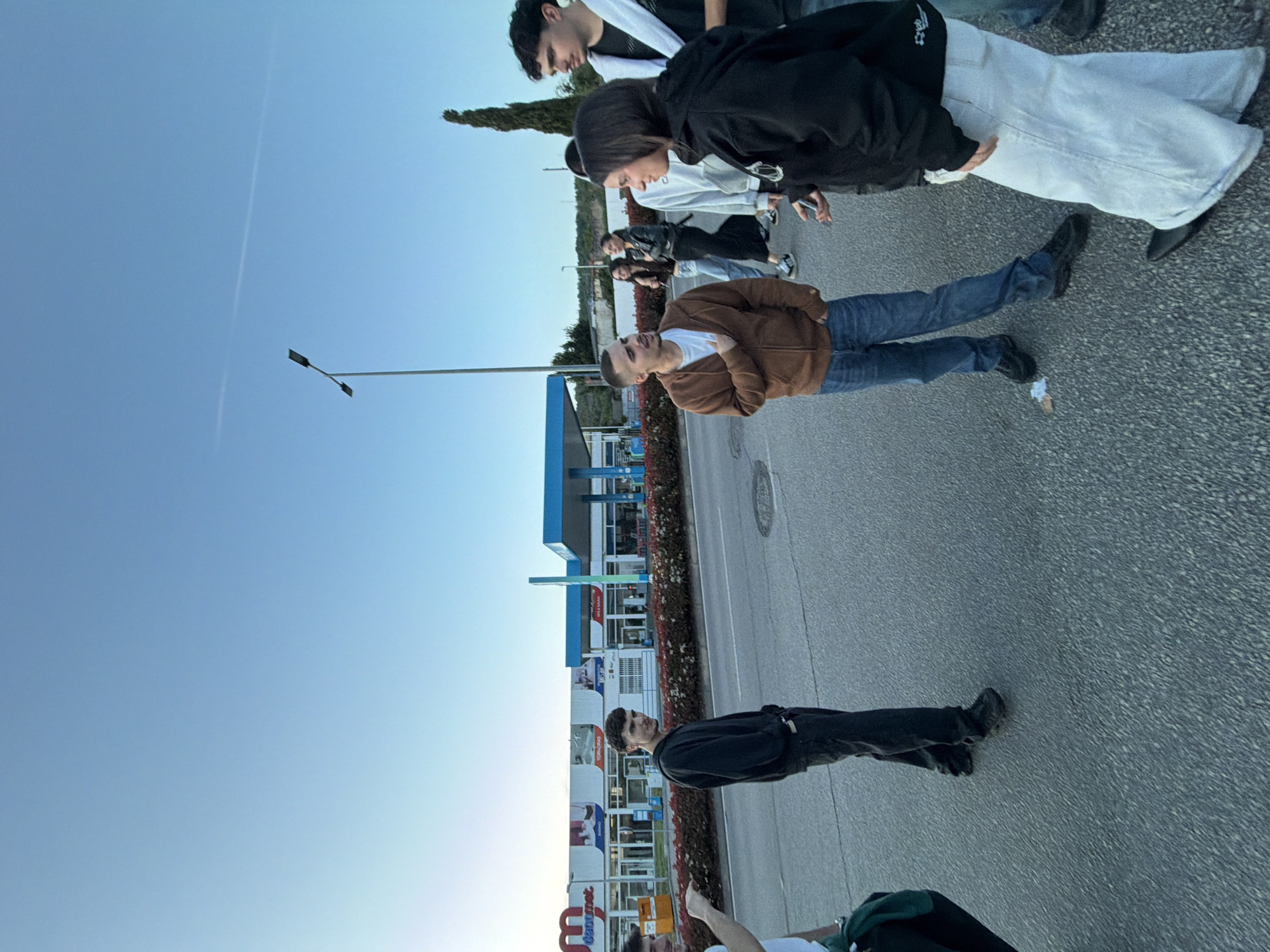 The image shows a group of people standing on an asphalt surface in Bragança, Portugal. The sky is light blue. There's a row of buildings behind the group, including a gas station and other commercial structures. A row of red flowers separates the pavement from the buildings. A street light is visible against the blue sky. The group consists of individuals of varying ages. One person is wearing a brown jacket and blue jeans. Another wears a black jacket and pants. Some are dressed in white pants and black jackets. They appear to be walking or standing near the pavement.