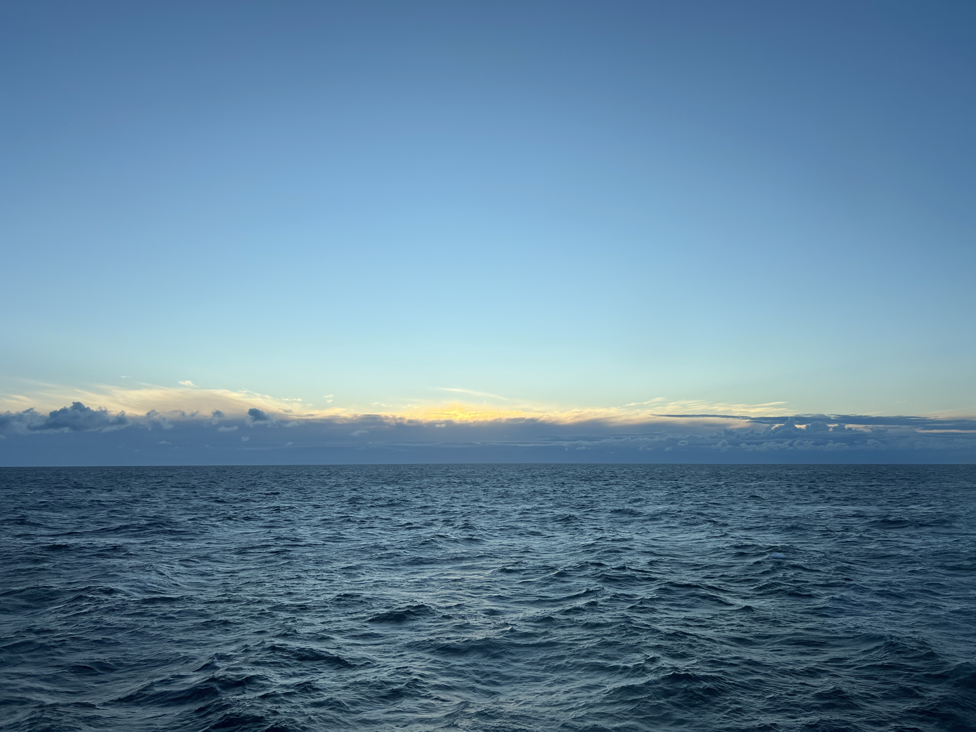 A wide shot captures a tranquil ocean under a vast, clear blue sky. The sun is setting behind a band of clouds on the horizon, casting a soft yellow glow. The ocean's surface is gently rippled, reflecting the colors of the sky. There are no people or structures visible in the image. The setting appears to be a natural, coastal environment. The location in the city of Unknown, Poland is not discernible from the image.