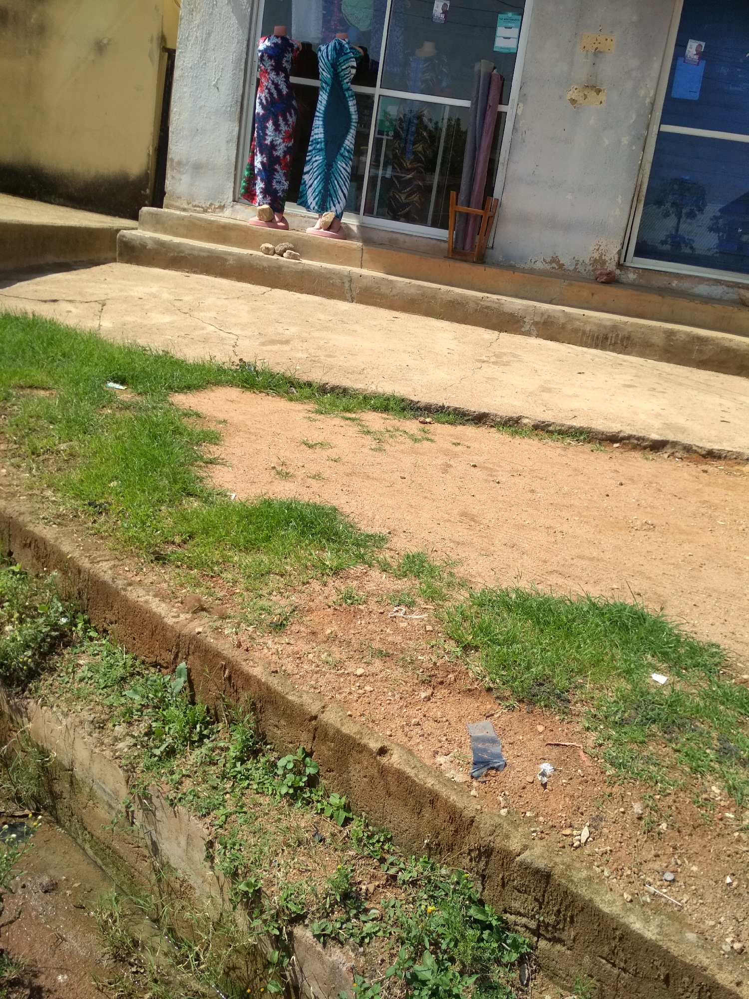 This image captures an outdoor scene, likely a street or pathway in a commercial area of Jos, Nigeria, under bright daylight. The foreground features a rough, sandy-brown dirt ground with scattered patches of green grass and some small pieces of litter. A concrete curb runs horizontally across the bottom, bordering a concrete ditch or drain filled with murky water and overgrown with weeds and grass.
In the midground, a set of concrete steps leads up to a building that appears to house a shop. Two headless mannequins are prominently displayed on the top step, each wearing a long, patterned dress. The mannequin on the left wears a blue, red, and white dress with a floral or abstract pattern, while the one on the right is dressed in a teal and white tie-dye or batik-style garment. Both mannequins stand on pink circular bases, with small rocks placed in front of them on the steps, possibly to stabilize them.
The background consists of the shop's facade. It features large glass windows that reflect the sky and a nearby tree. Some small posters or flyers are affixed to the windows, but any text is illegible. The building's wall is a light, possibly off-white or gray color, showing some signs of wear and peeling paint in places. Leaning against the wall next to the window is a simple wooden frame holding several rolled-up items, possibly fabrics or mats. The clear, sunny weather casts distinct shadows, indicating a mid-day setting. No people are visible, and no specific activity or event is taking place beyond the display of merchandise.