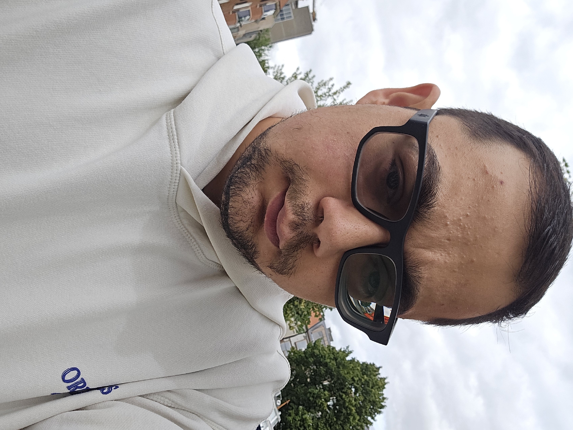 A medium shot captures a person wearing glasses and a white hooded sweatshirt, looking down slightly. The background is an overcast sky with some trees and buildings visible on the left side, suggesting an urban outdoor setting. The buildings appear to be typical of Ghent, Belgium, with some architectural elements visible. The person's face is the primary focus, with the details of their facial features and the clothing clearly visible. The overall mood is neutral and candid.